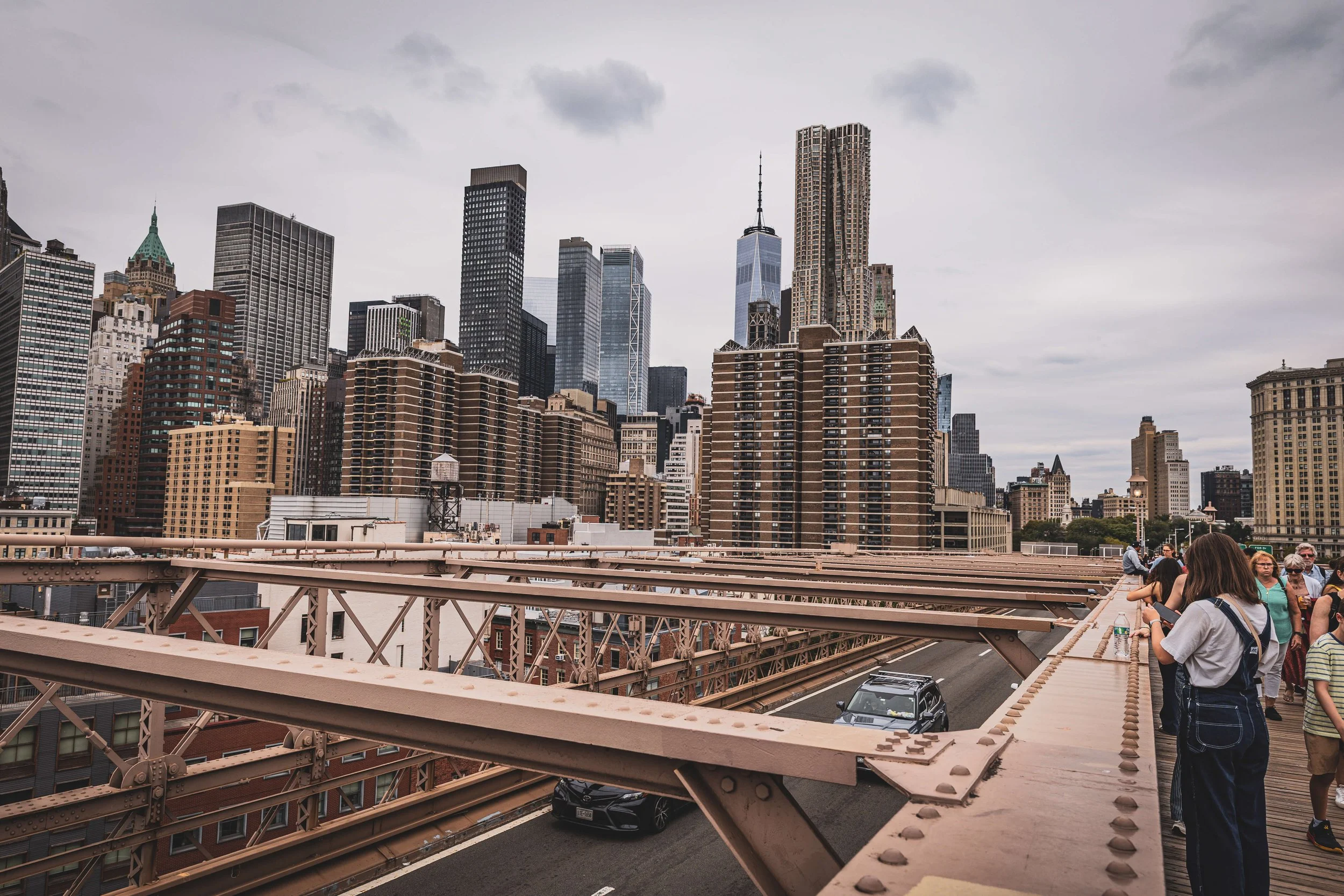 People stand on a viewing platform of the Brooklyn Bridge, overlooking Manhattan skyscrapers in New York City with cloudy sky.