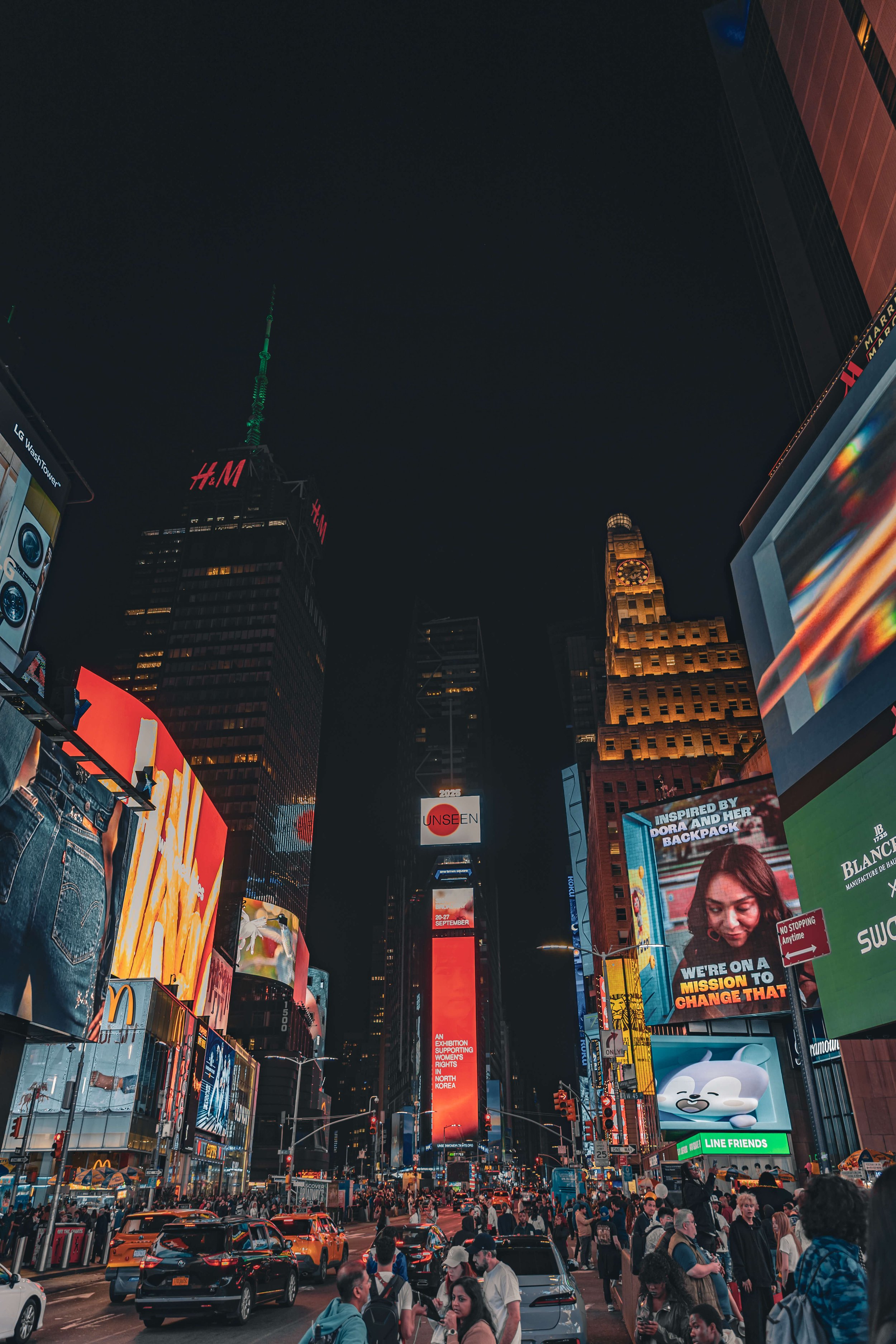 Nighttime scene in Times Square, New York City, showing tall buildings with bright electronic billboards, a crowd of pedestrians, and vehicles on the street.
