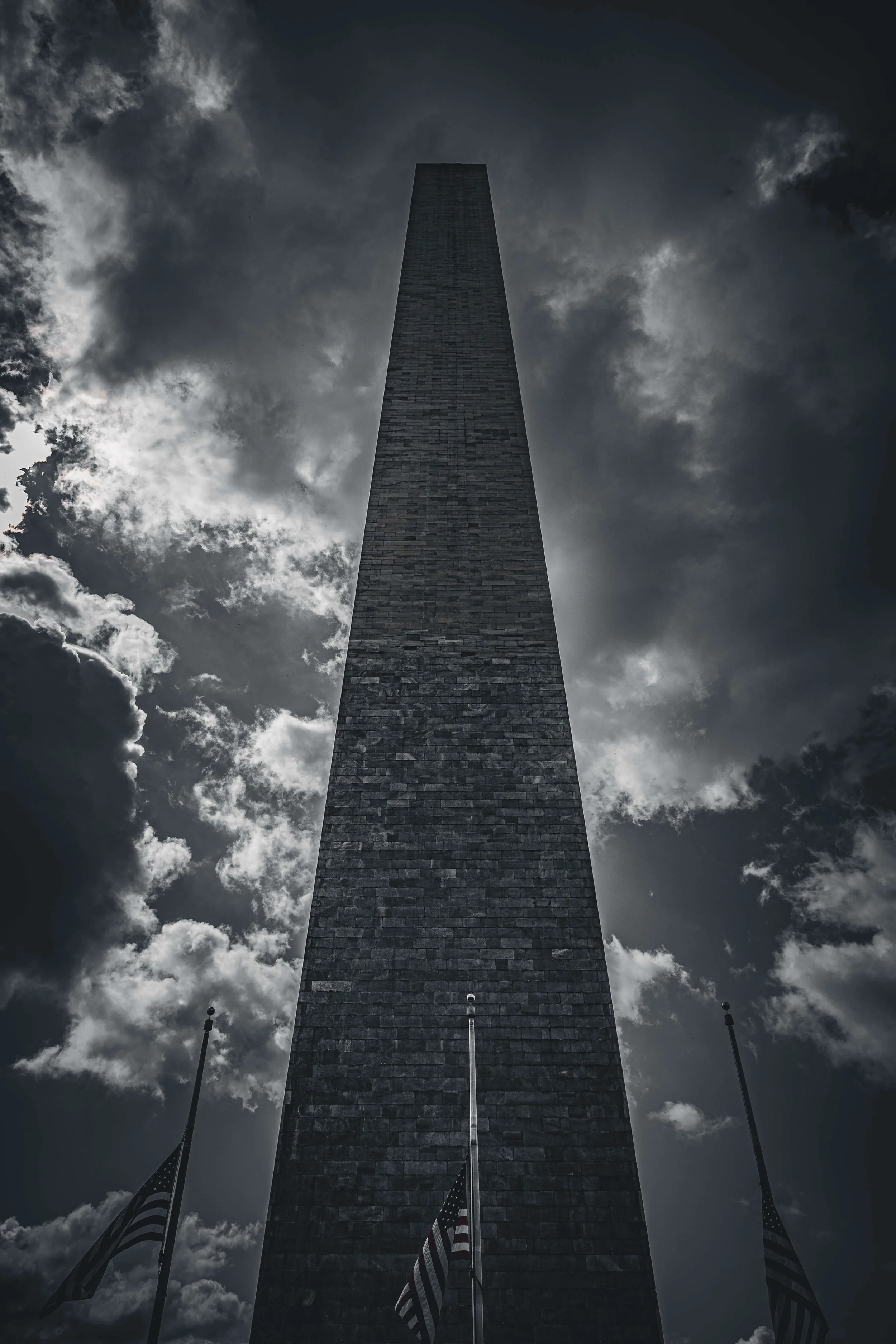 A tall, dark monument viewed from below, with cloudy skies and flags at the base.