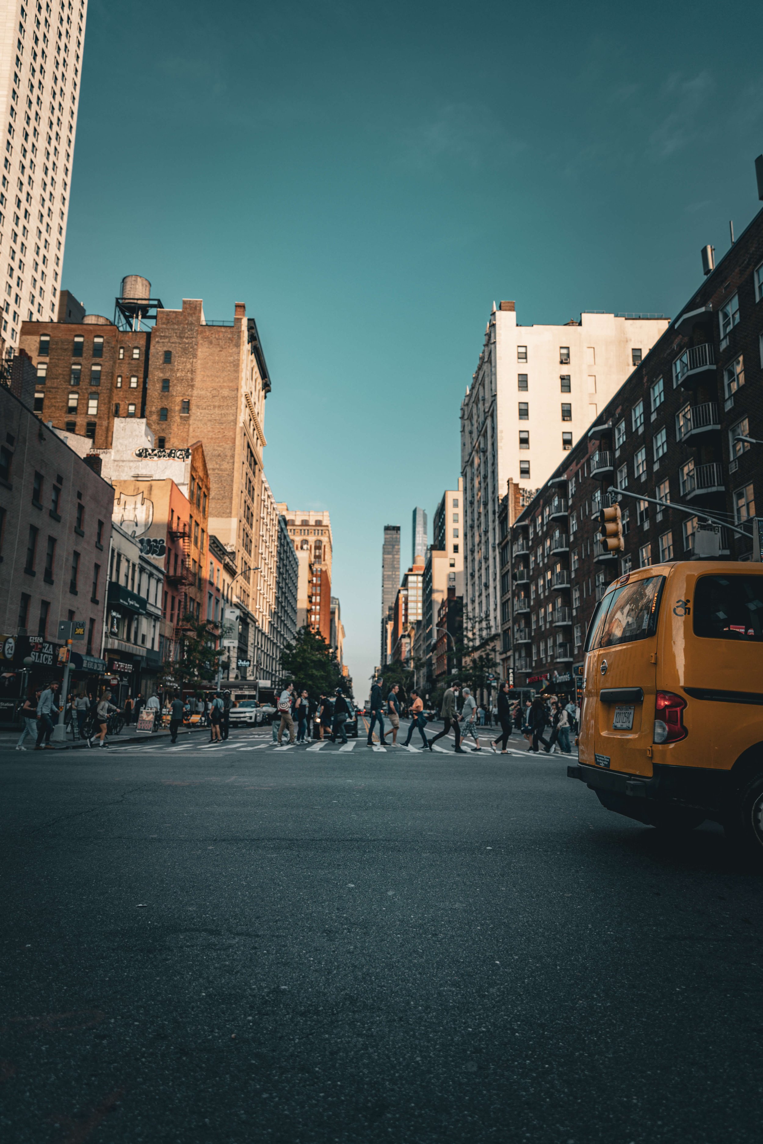 City street scene with tall buildings, pedestrians crossing the street, and a yellow taxi cab on the right side.
