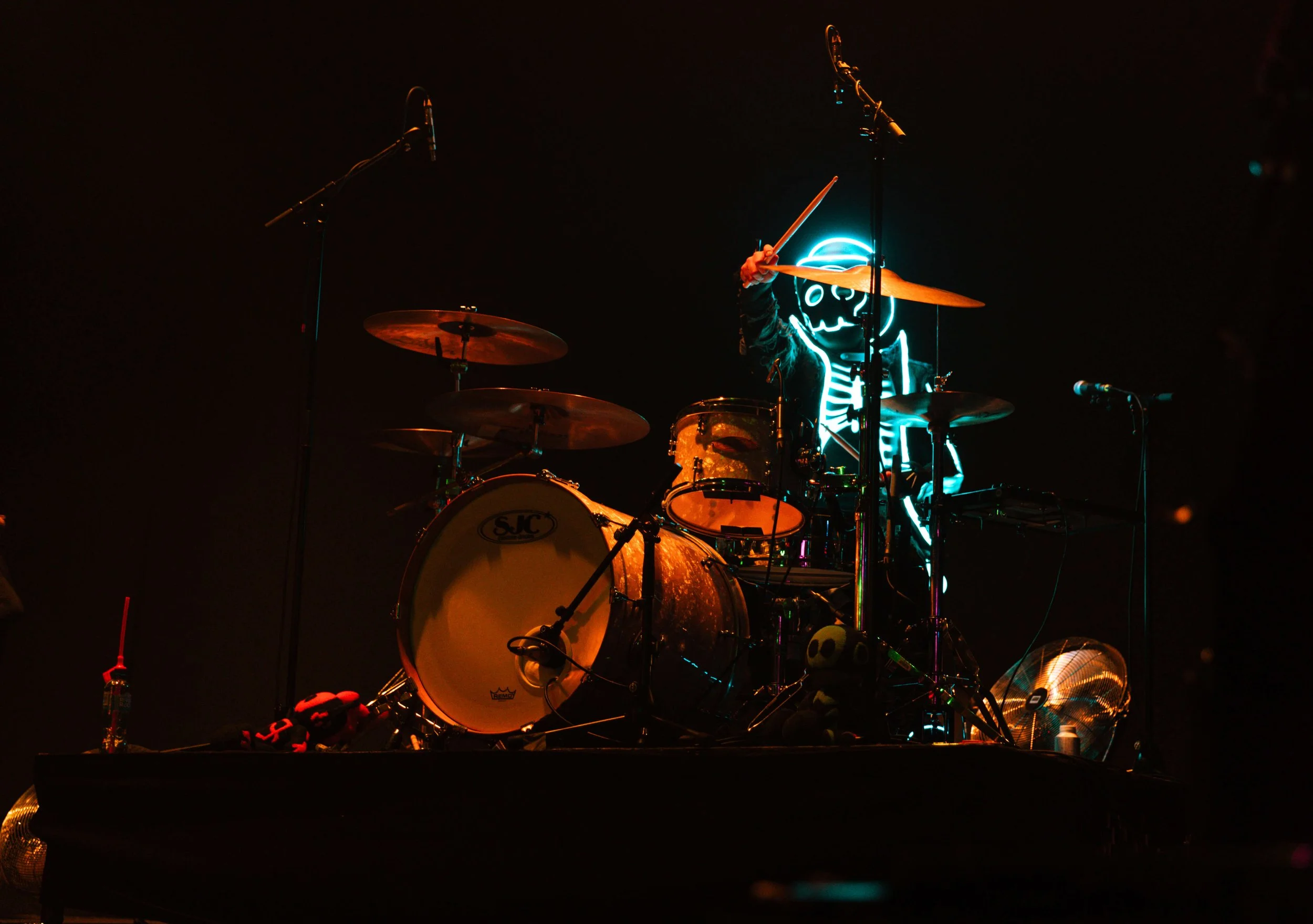 A drum set on stage with a neon skeleton wearing a hat glowing in the background, illuminated by stage lights, with microphones and other musical equipment around.