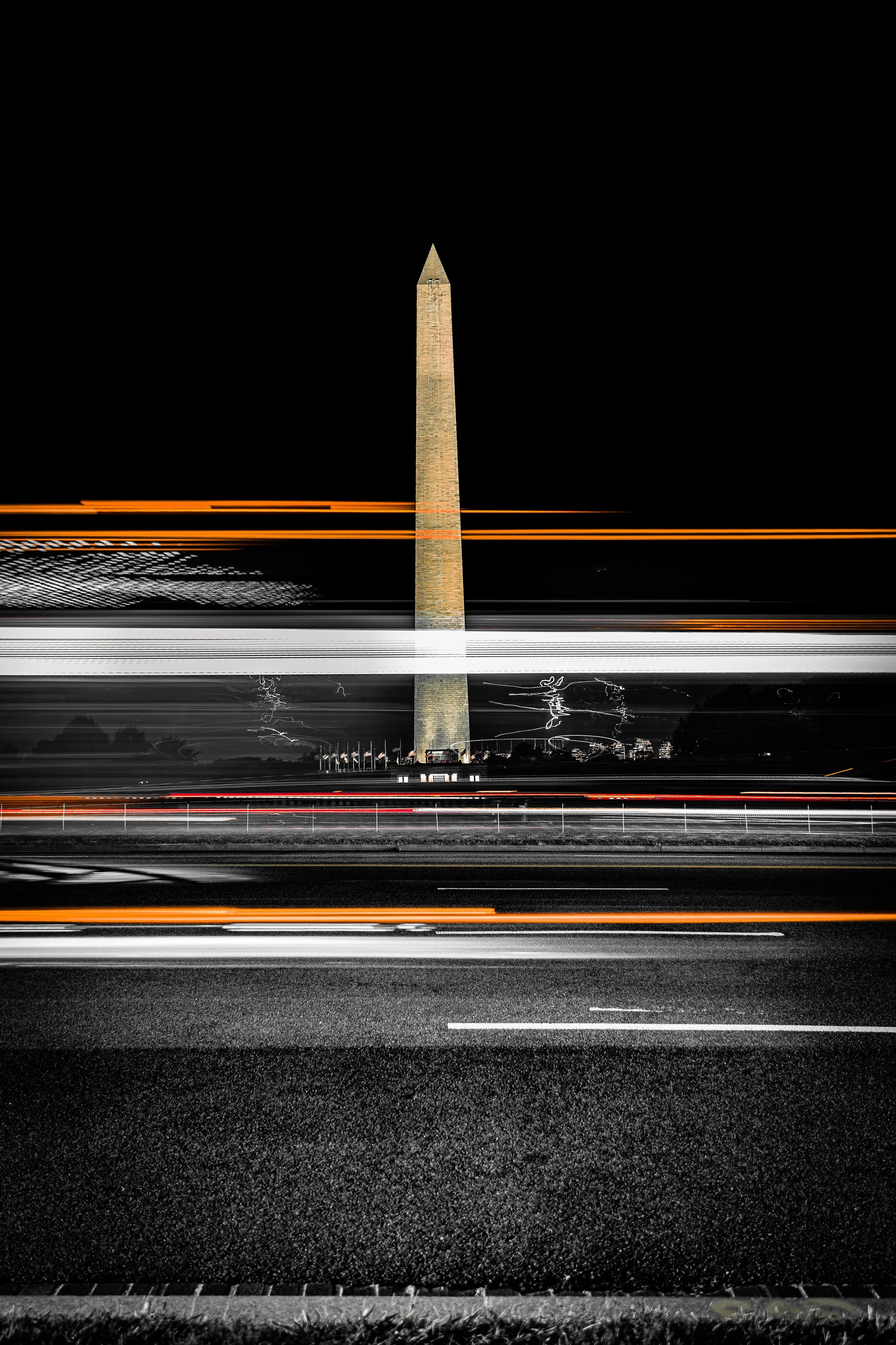 Nighttime view of the Washington Monument illuminated, with light trails from moving vehicles across a highway in the foreground.