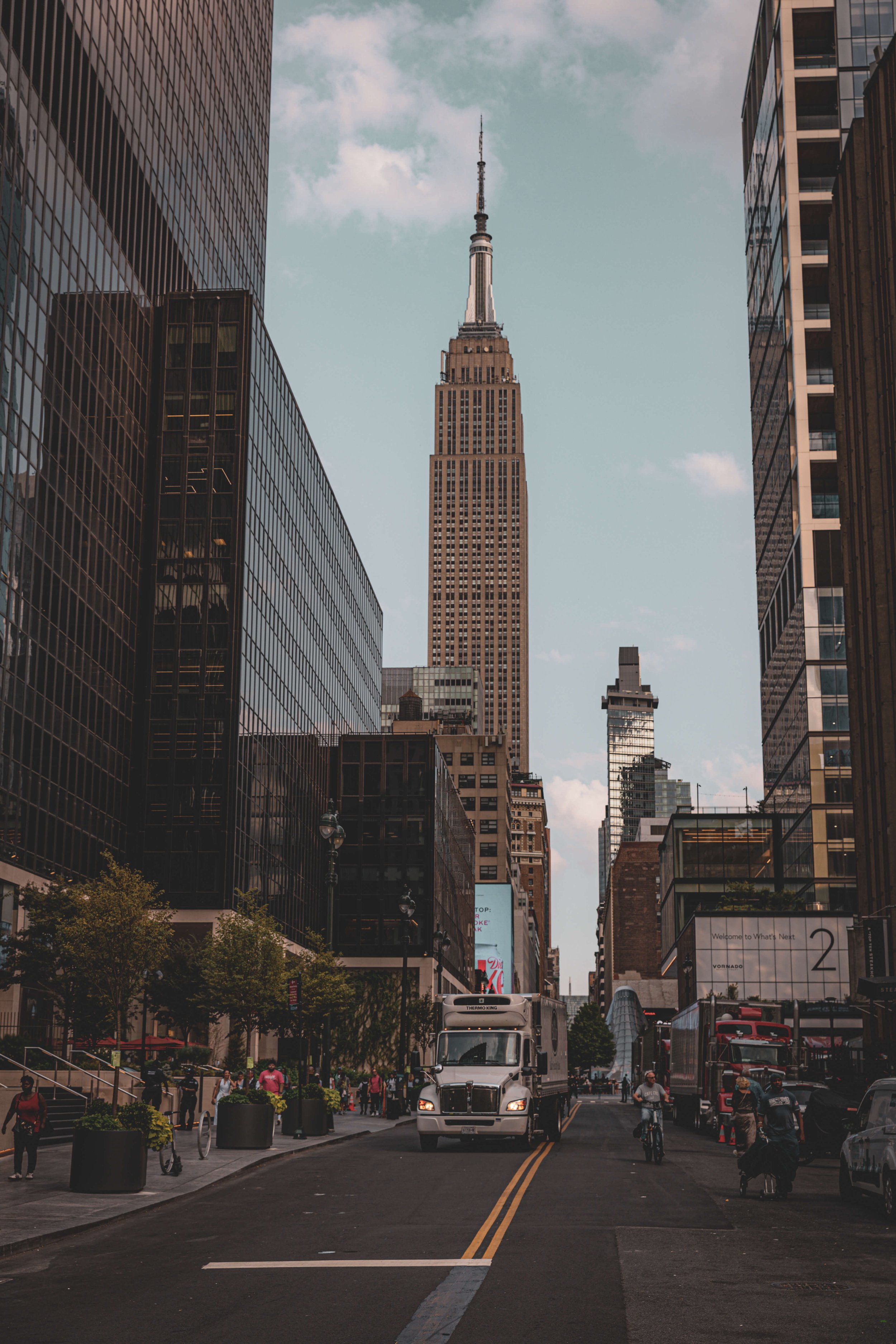 New York City street scene with the Empire State Building towering in the background, surrounded by modern skyscrapers, with traffic, pedestrians, and trees on the street.