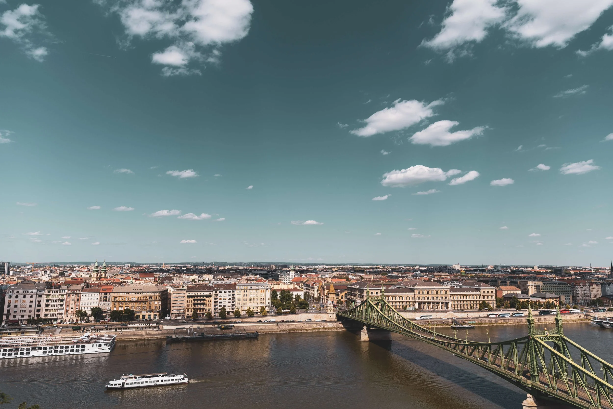 A cityscape view of Budapest, Hungary, featuring the Danube River with a cruise ship, a historic green bridge, and a skyline of historic buildings under a partly cloudy sky.