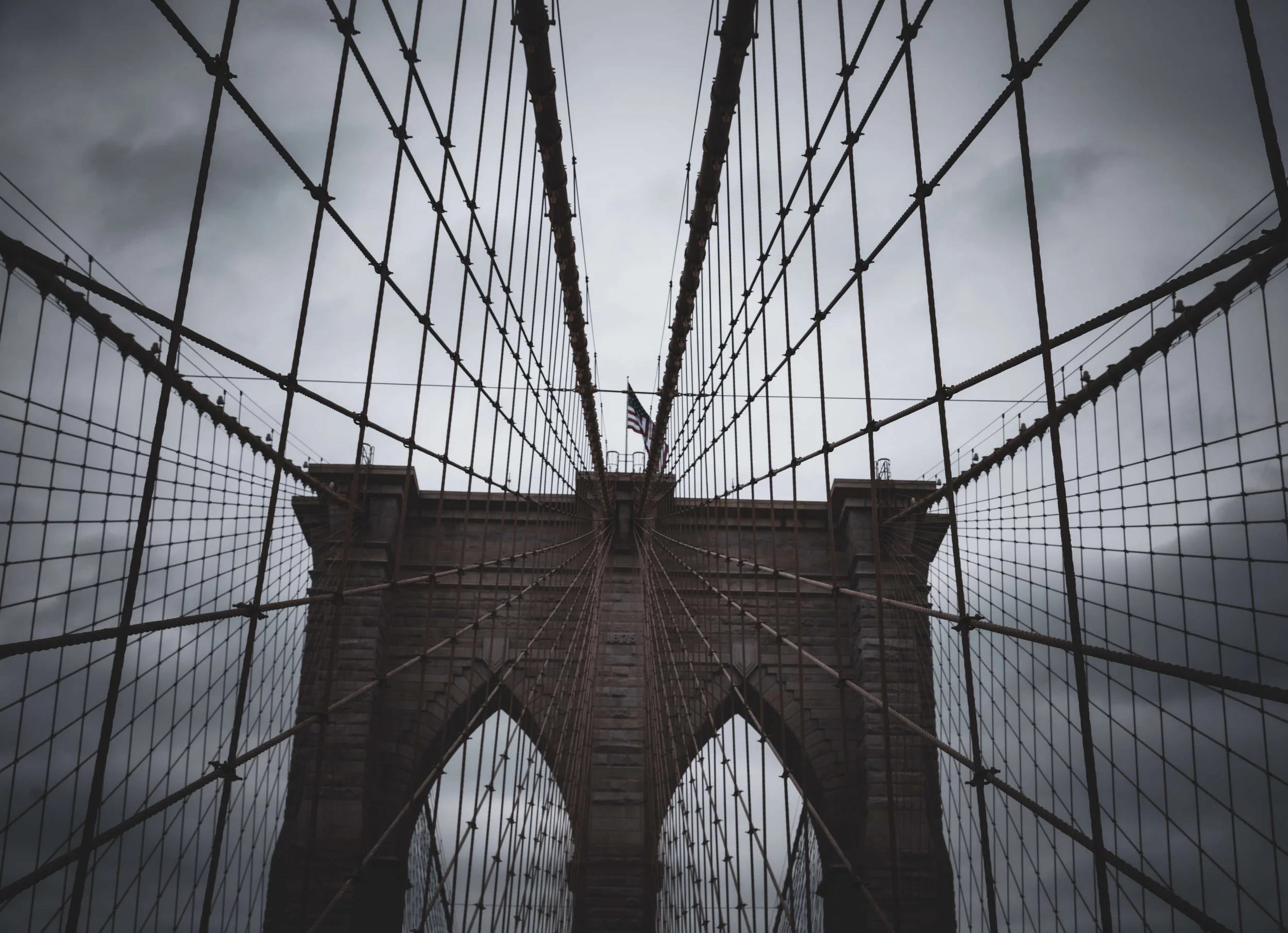Looking up at the Brooklyn Bridge's stone towers and steel cables against a dark, cloudy sky with an American flag at the top.