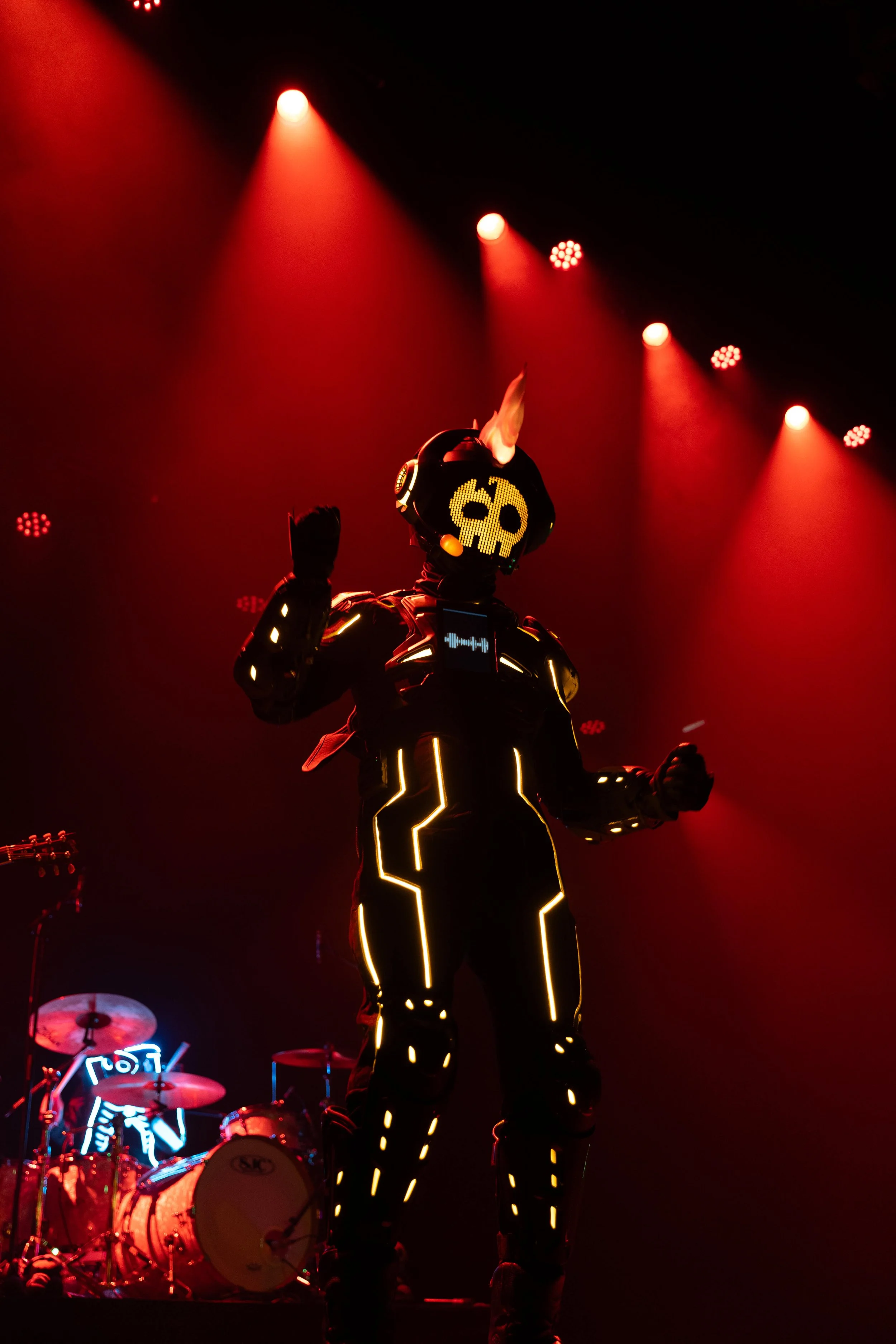 Performer dressed in glowing, futuristic LED costume with skull design, on stage under red spotlights, with drums in the background.