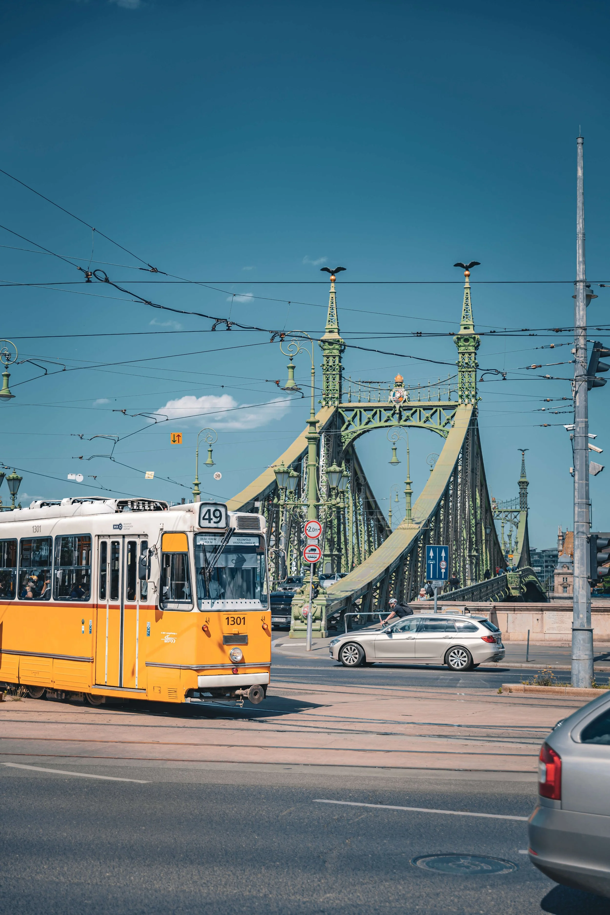 A street scene with a yellow tram, cars, and a bridge with towers topped by bird-shaped spires, under a clear blue sky.