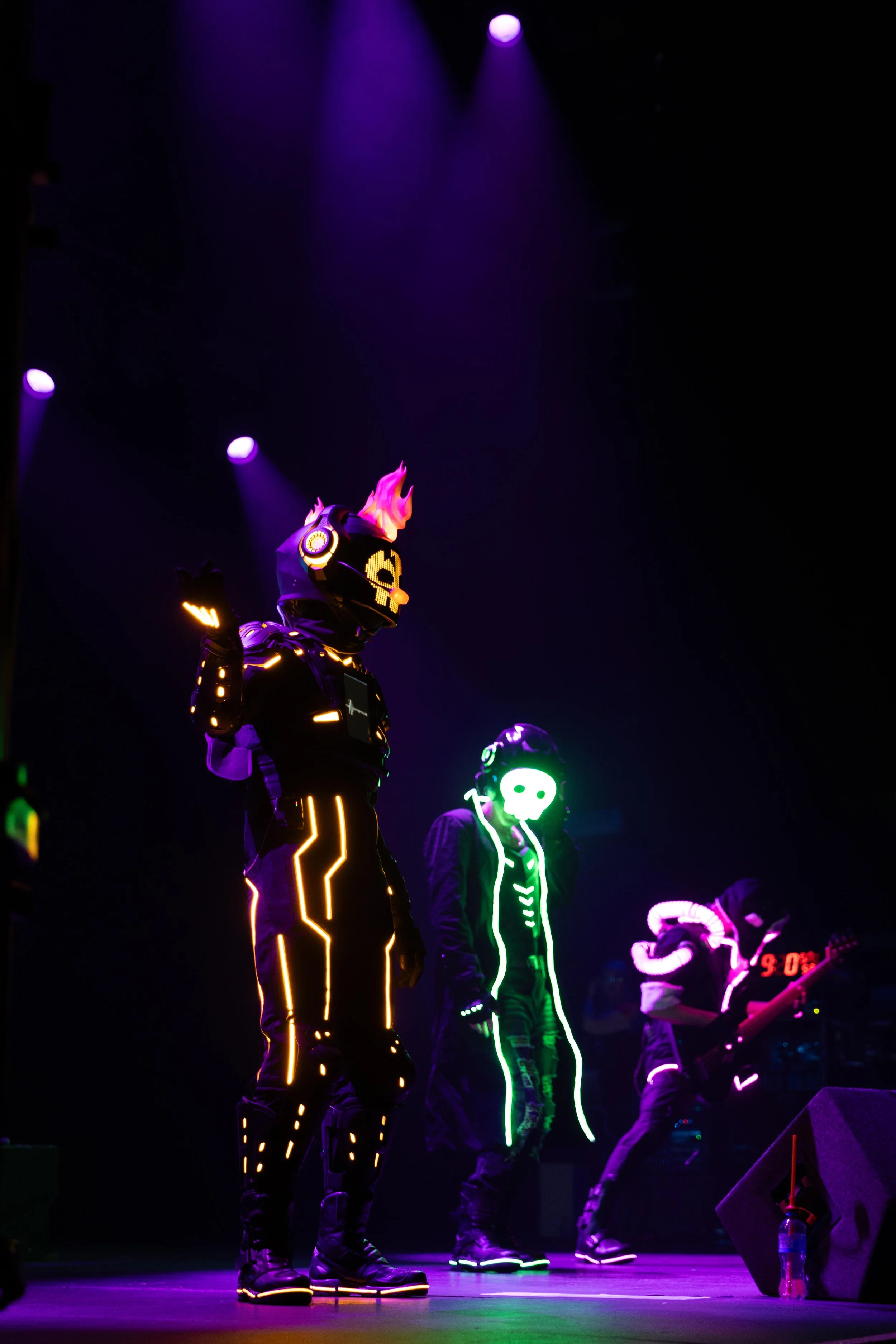 Performers on stage dressed in black with glowing, colorful LED costumes, including one with flaming headgear, illuminated lines on clothing, purple lighting overhead, and a musician in the background.