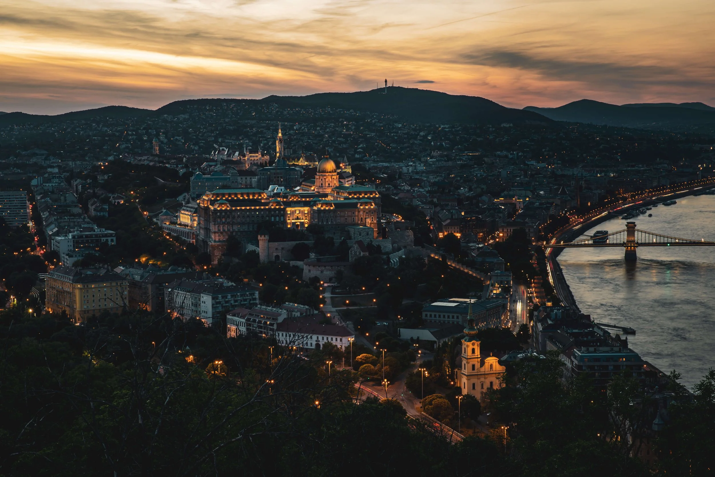 A city skyline at dusk with illuminated buildings, a river along the city edge, and a bridge. Hills are visible in the background with a sunset sky.