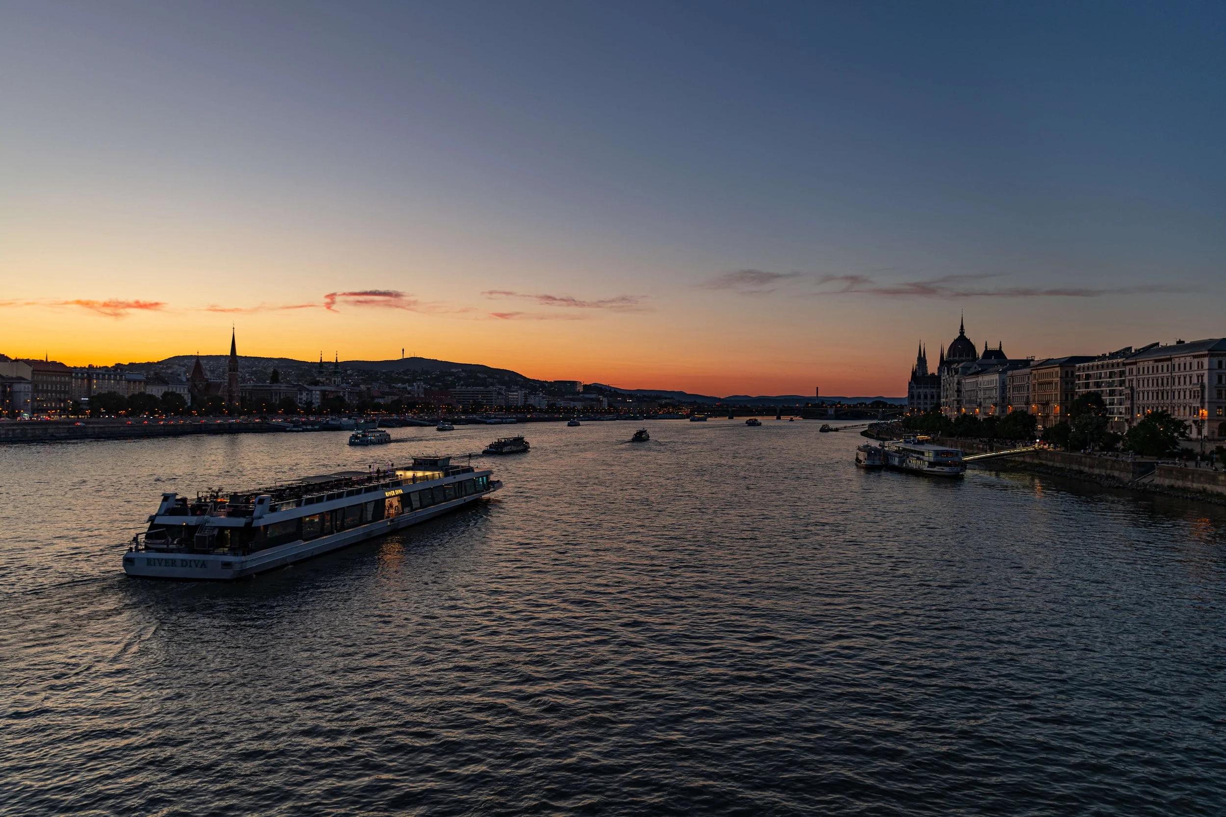 Sunset over a river with boats and historic European buildings along the riverbank.