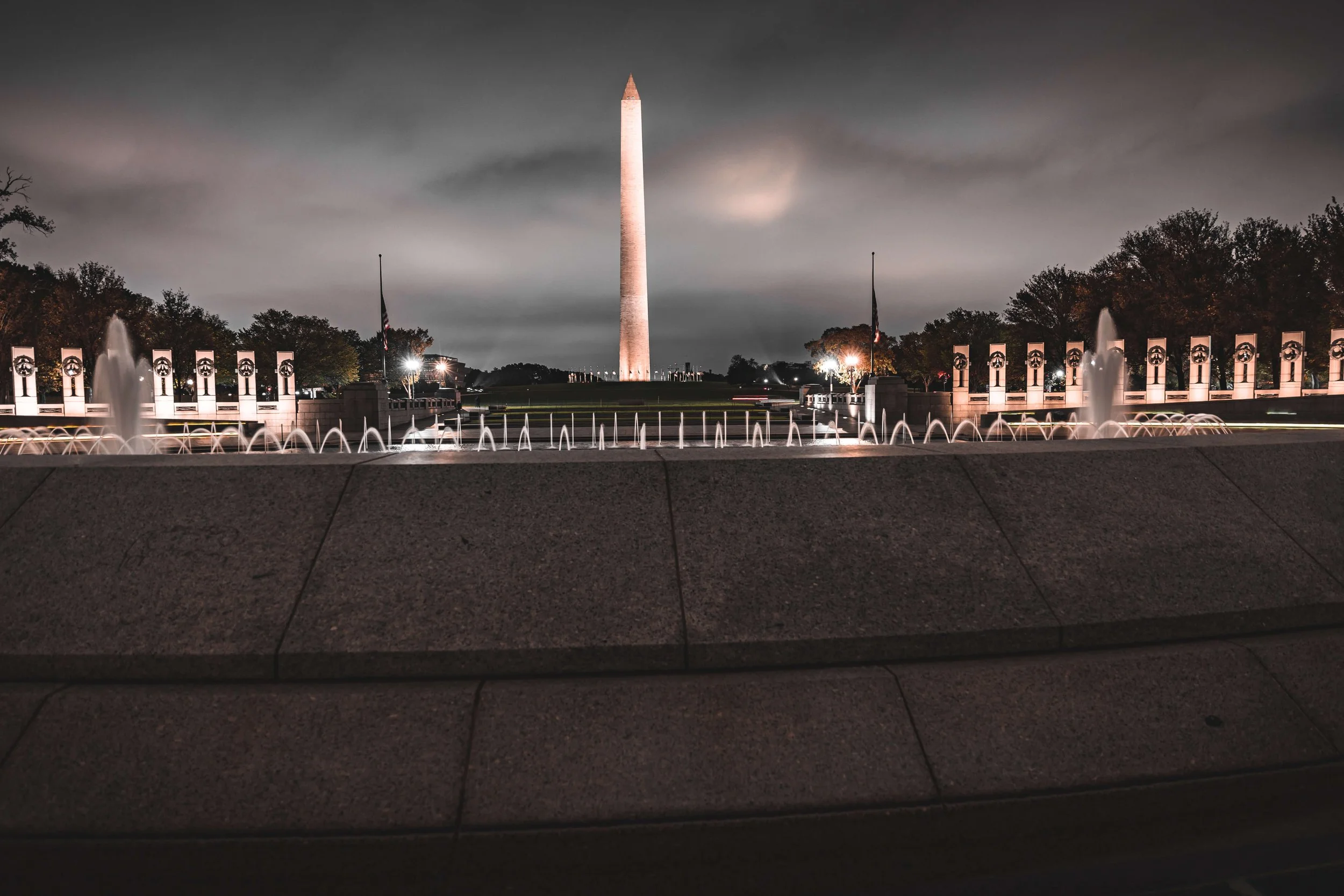 Night view of the Washington Monument in Washington, D.C., with illuminated fountains in the foreground and flags on either side.
