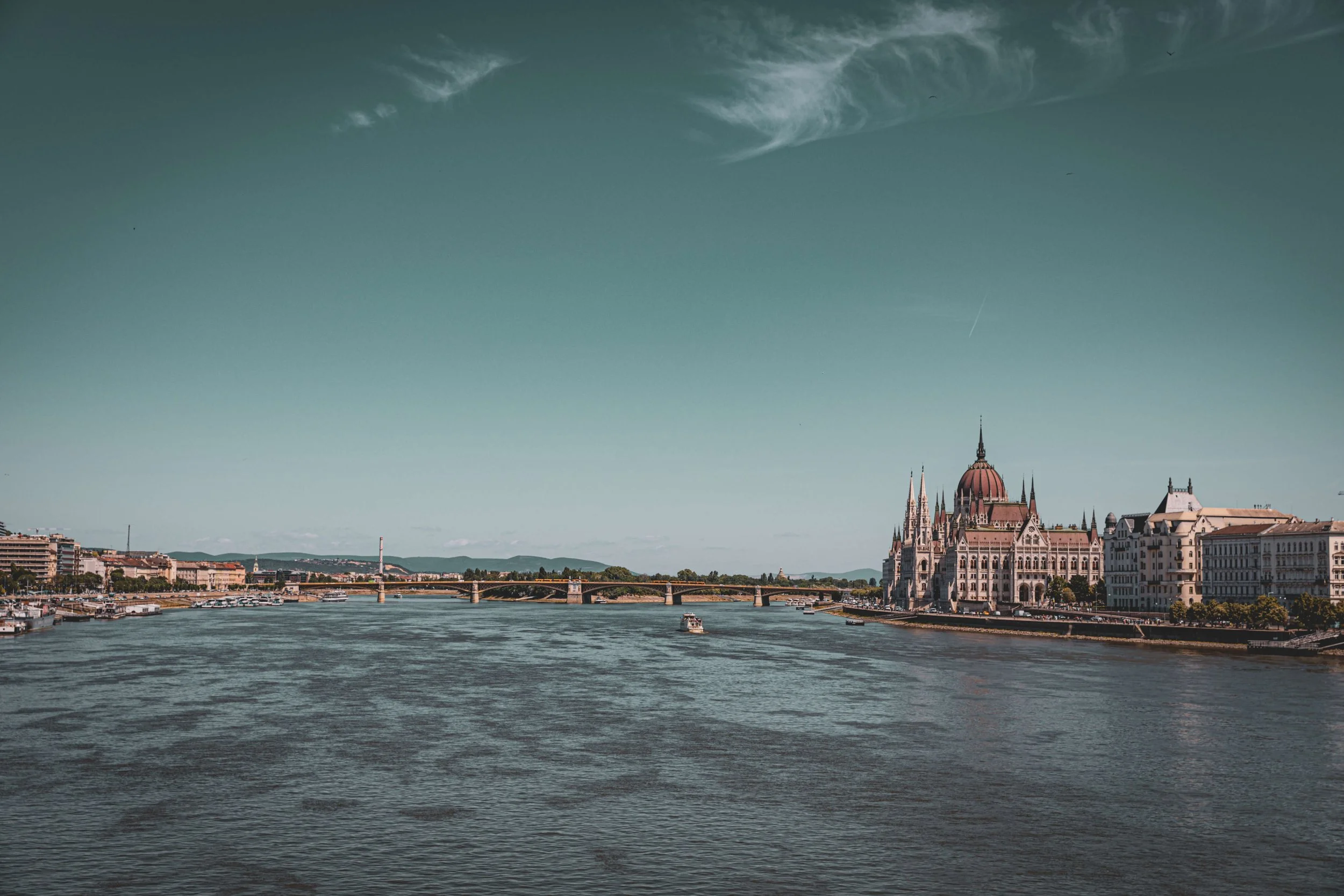View of the Danube River with boats and a historic Hungarian Parliament building in Budapest, Hungary, under a partly cloudy sky.