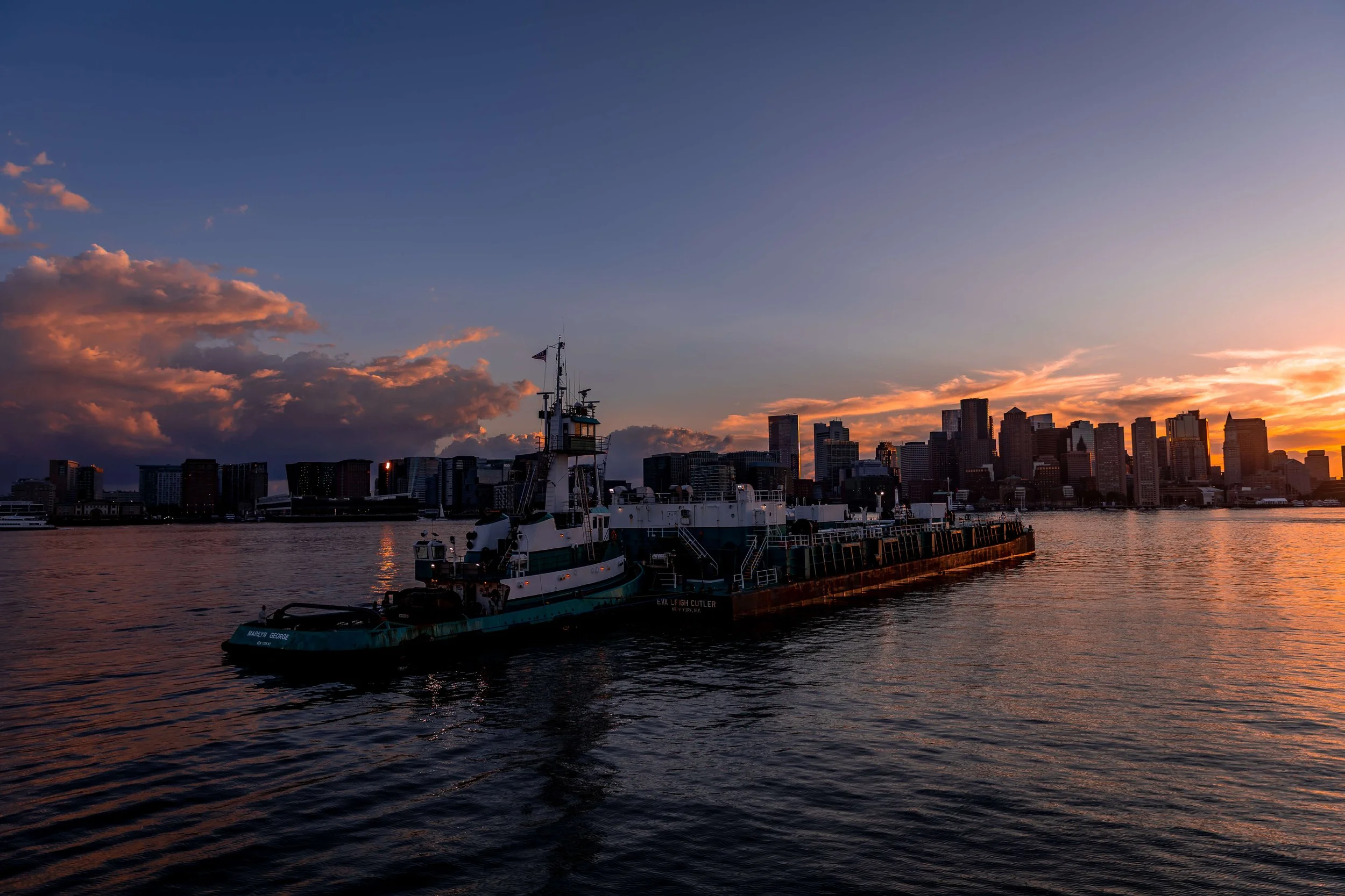 A tugboat on calm water with a city skyline at sunset in the background.