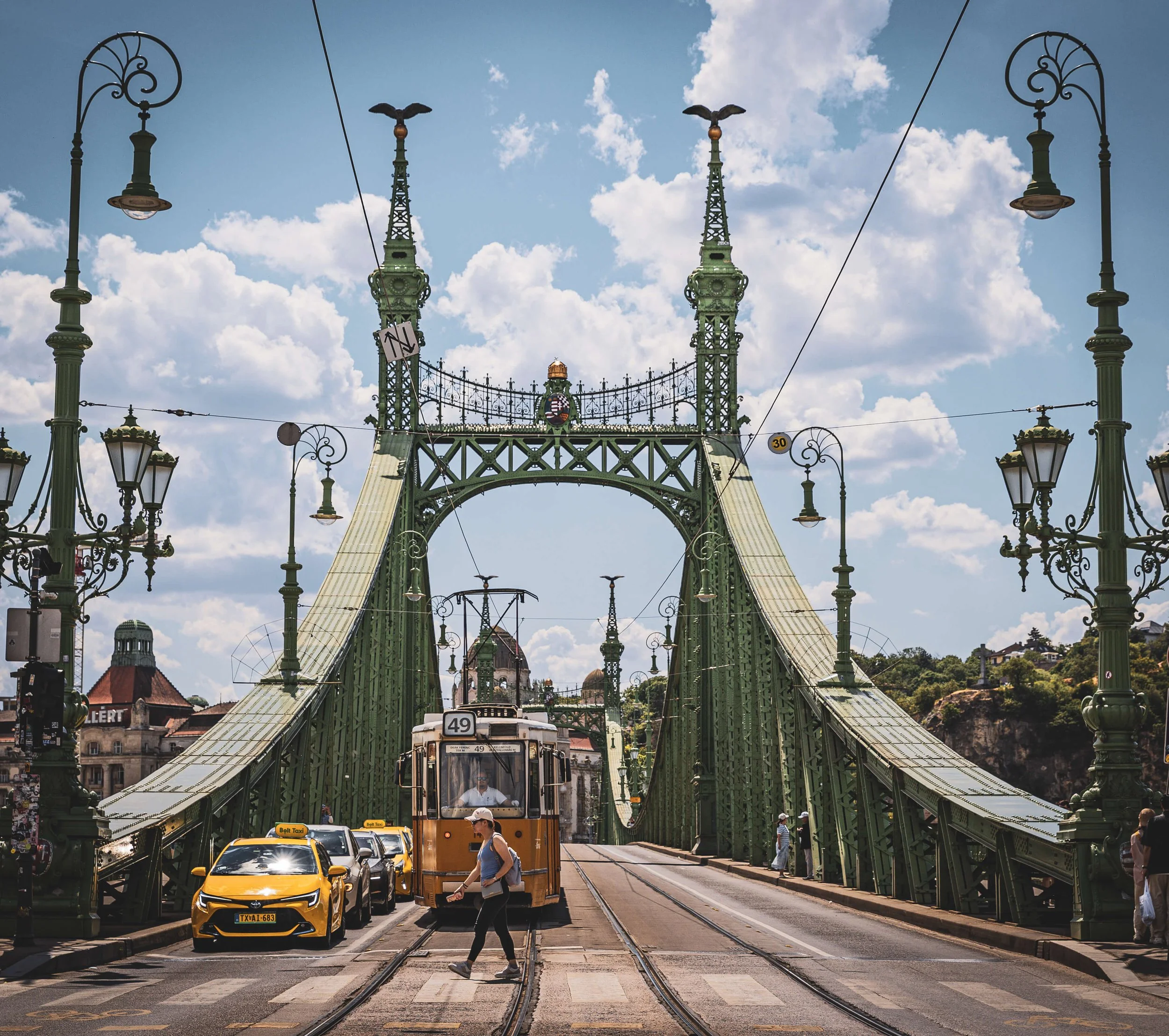 A busy city street scene featuring a bridge with ornate green metalwork and street lamps, with cars, a tram, pedestrians, and buildings under a partly cloudy sky.