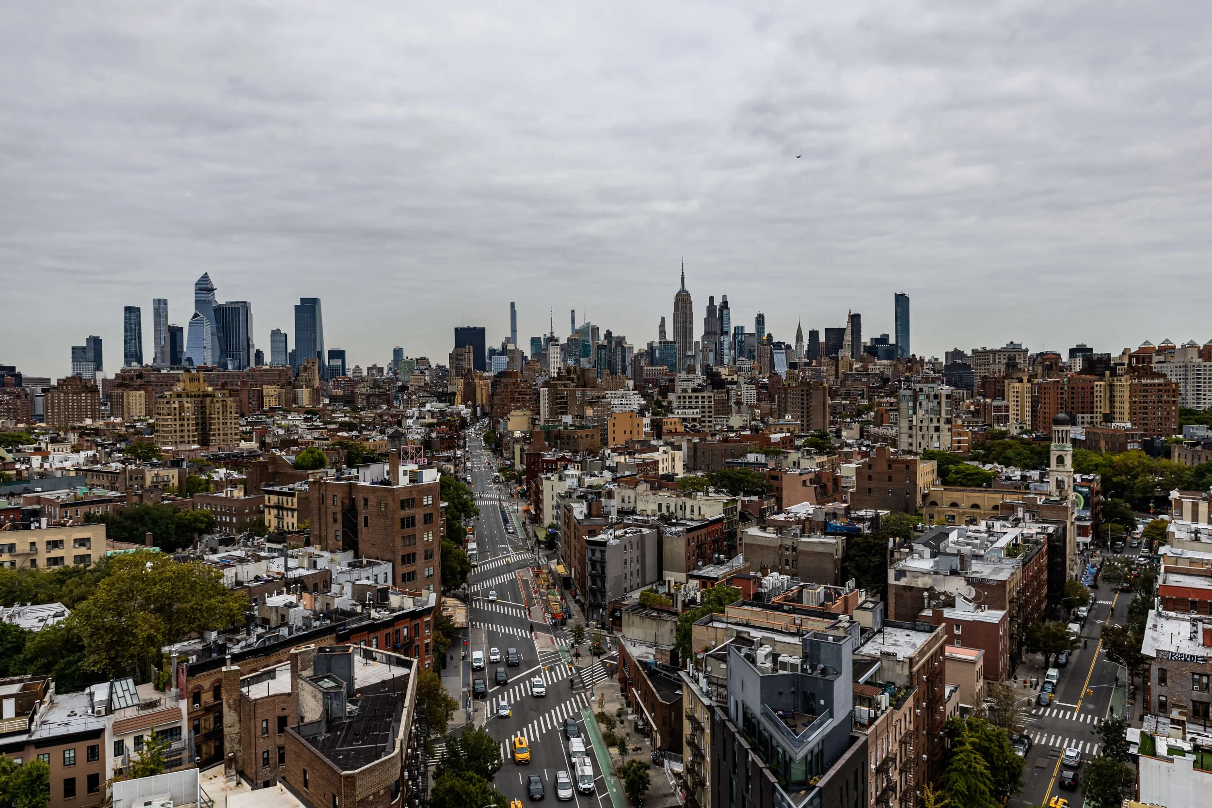 A panoramic view of New York City skyline with tall skyscrapers, some cloudy sky, busy streets, and buildings in the foreground.