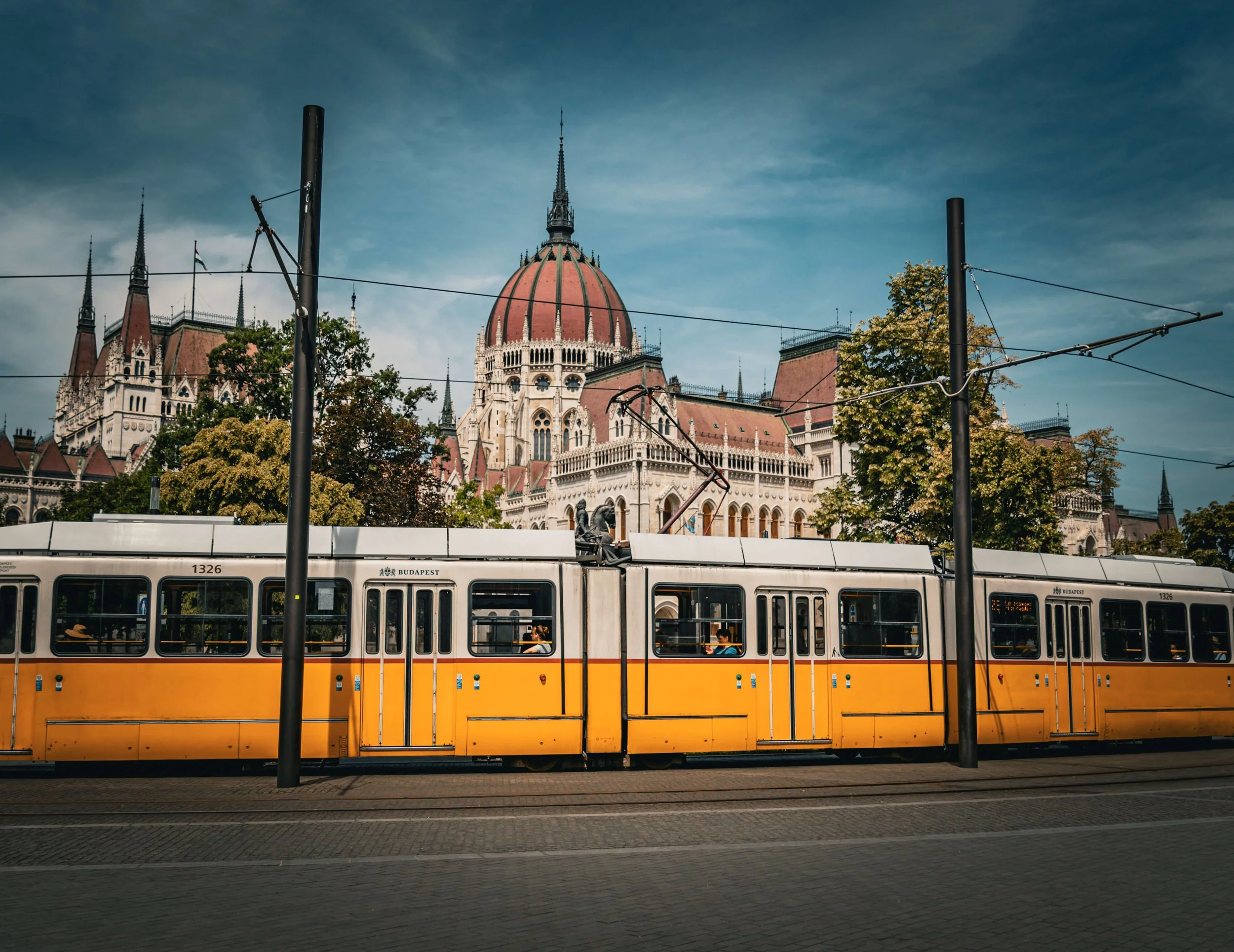 A yellow tram passes in front of a historic building with a large dome in Budapest, Hungary, under a partly cloudy sky.