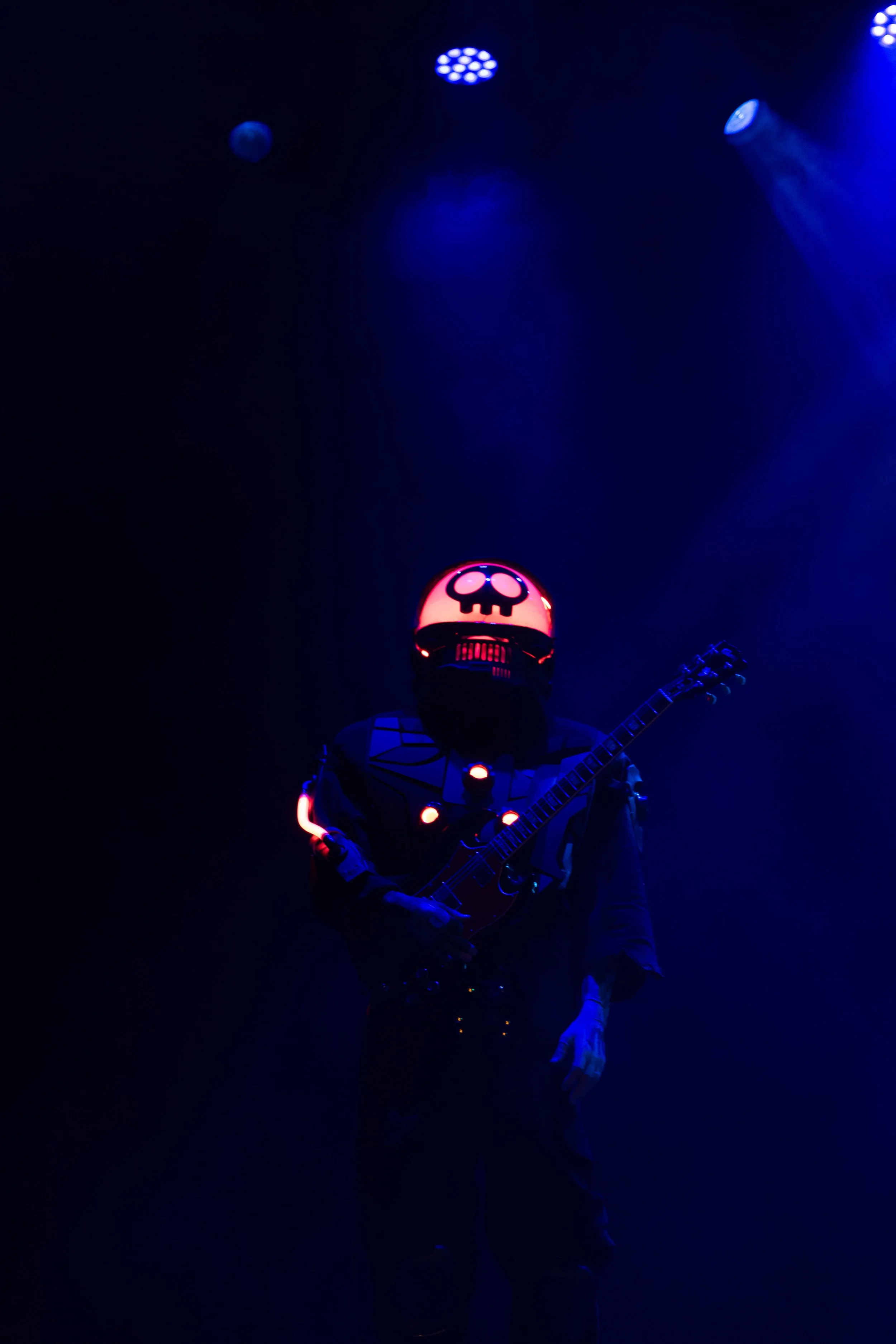 A performer wearing a illuminated skull mask and dark clothing, playing an electric guitar on stage with blue stage lighting and spotlight effects.