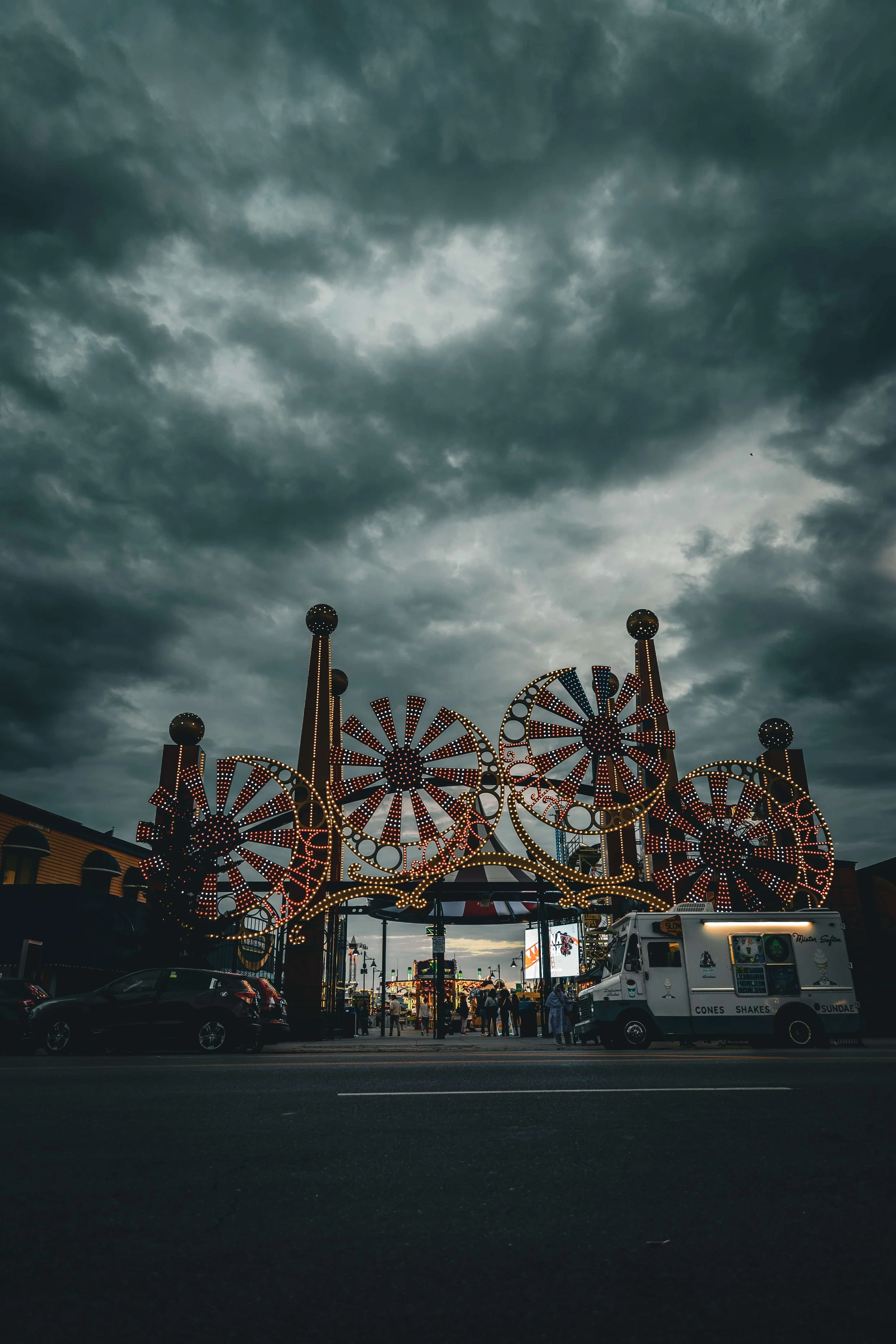 Nighttime scene of a fairground with illuminated Ferris wheels, food trucks, and people gathering, under dark stormy clouds in the sky.