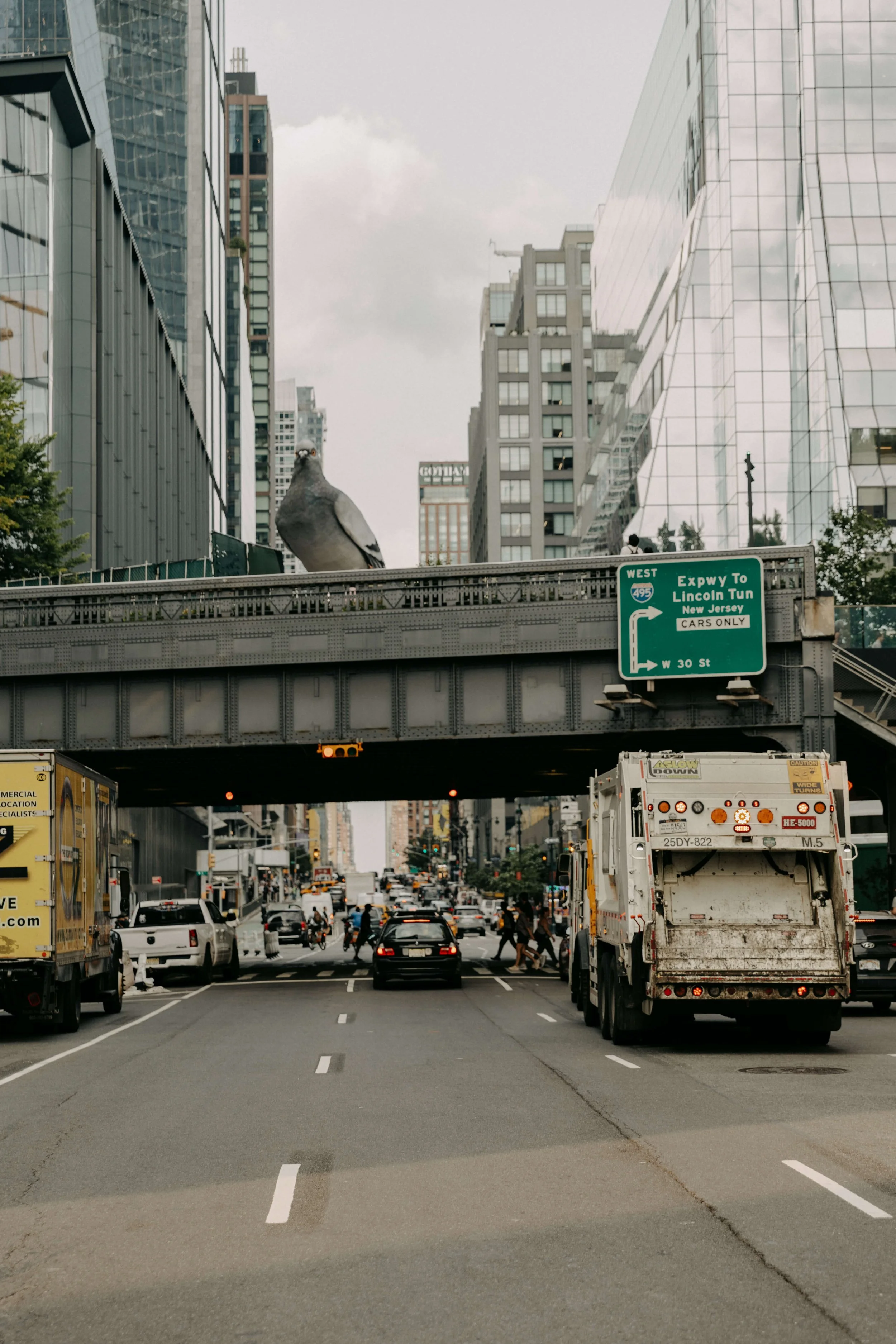 Street scene with vehicles, pedestrians, and a bridge with a large pigeon on top in an urban area with tall office buildings and highway signs.