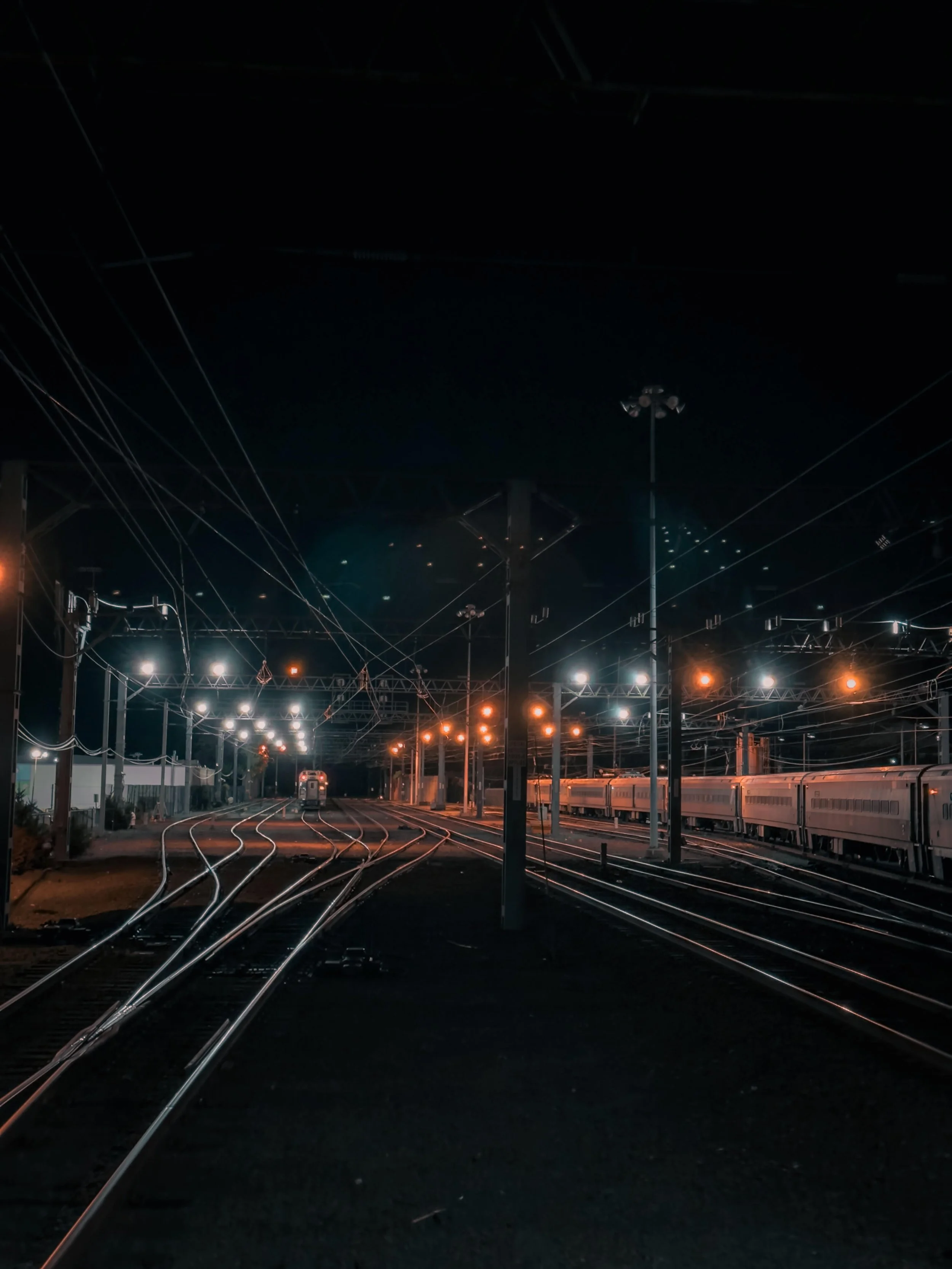 Nighttime scene of train tracks illuminated by streetlights with multiple train cars on the right and a train coming toward the viewer in the distance.