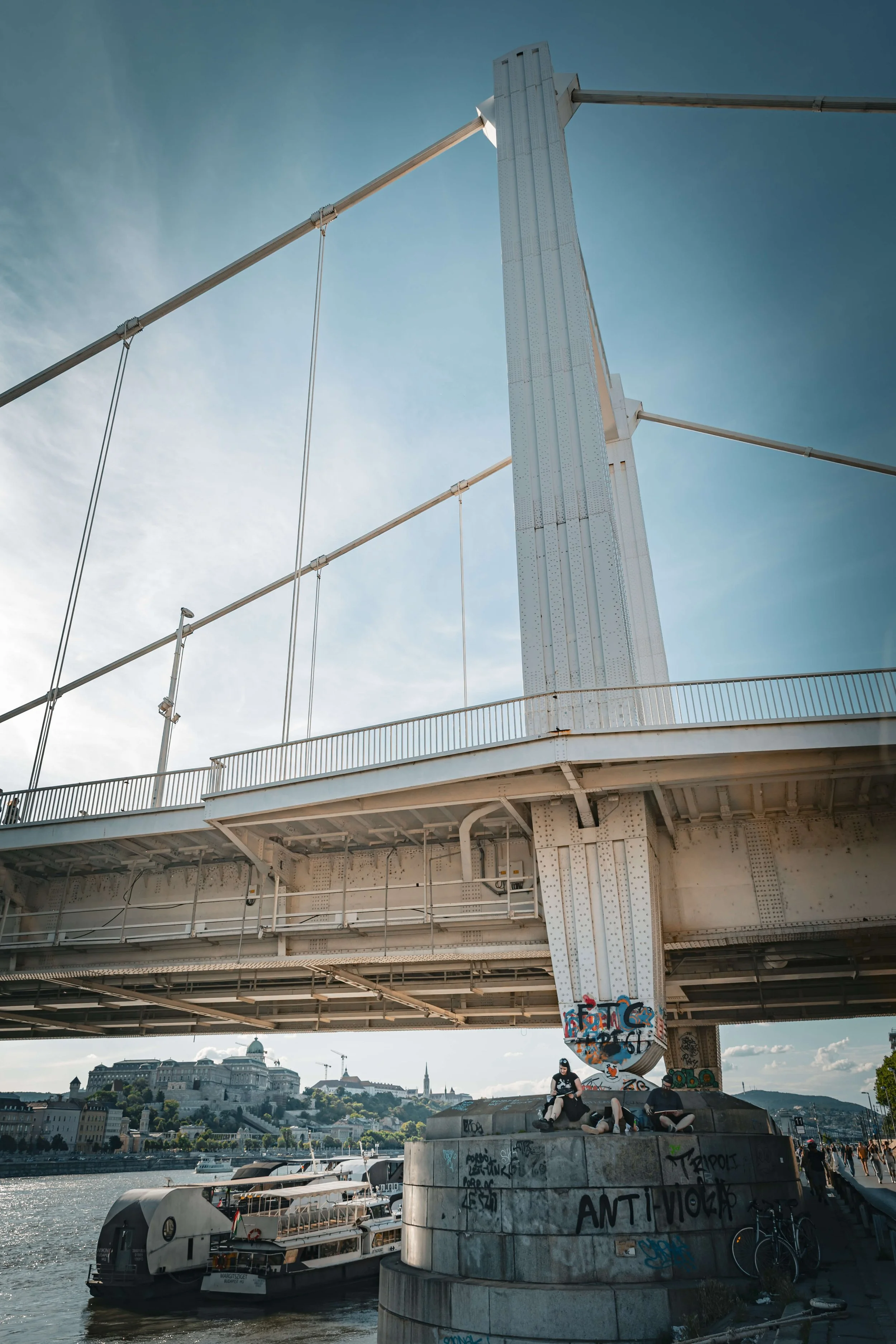 People sitting on a concrete platform under a large, white bridge with graffiti on the pillar, with a river and boats below, and a cityscape with historic buildings and a hill in the background.
