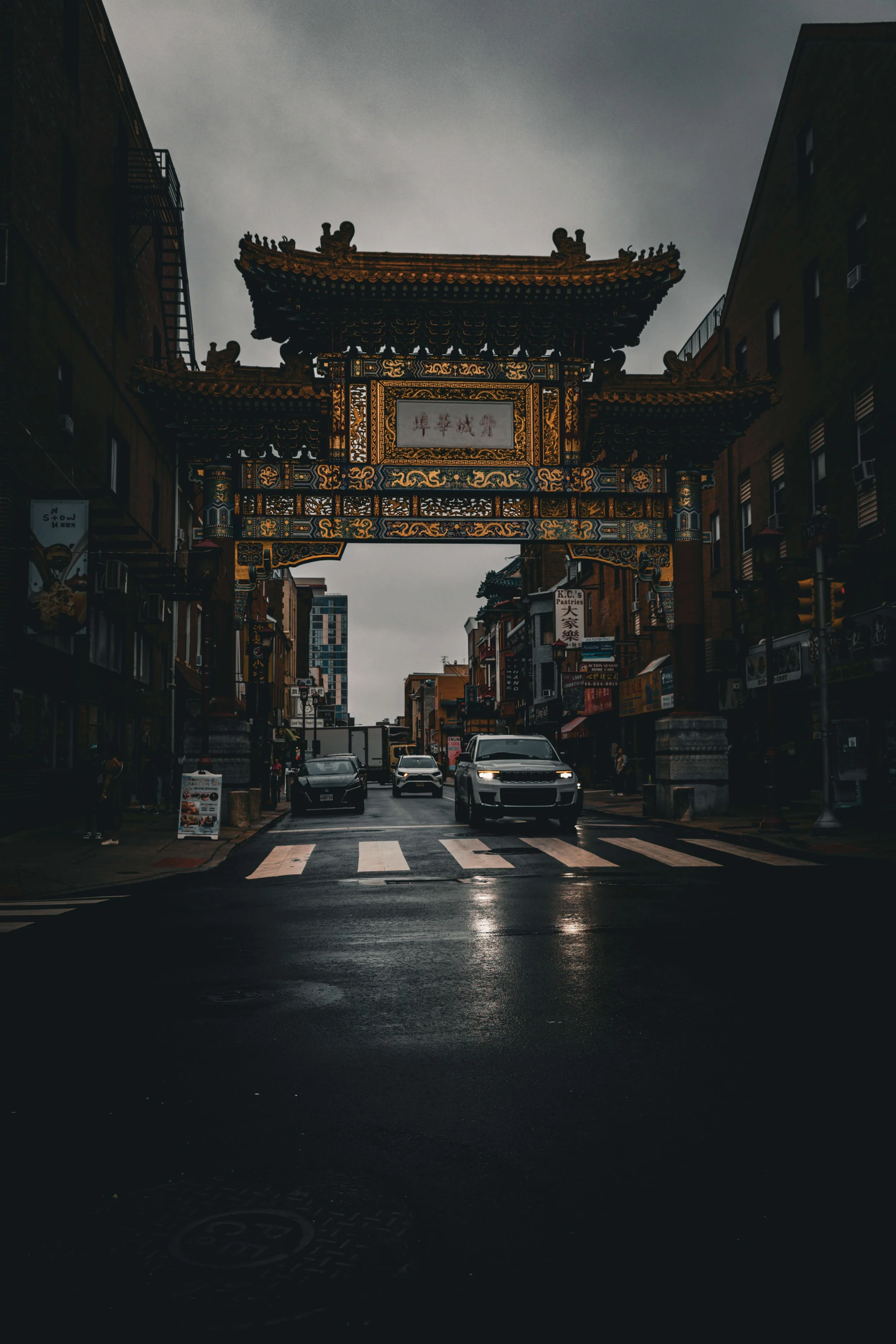 Street scene featuring an ornate Asian-style gateway with traditional Chinese architectural details, situated among city buildings on a rainy day.