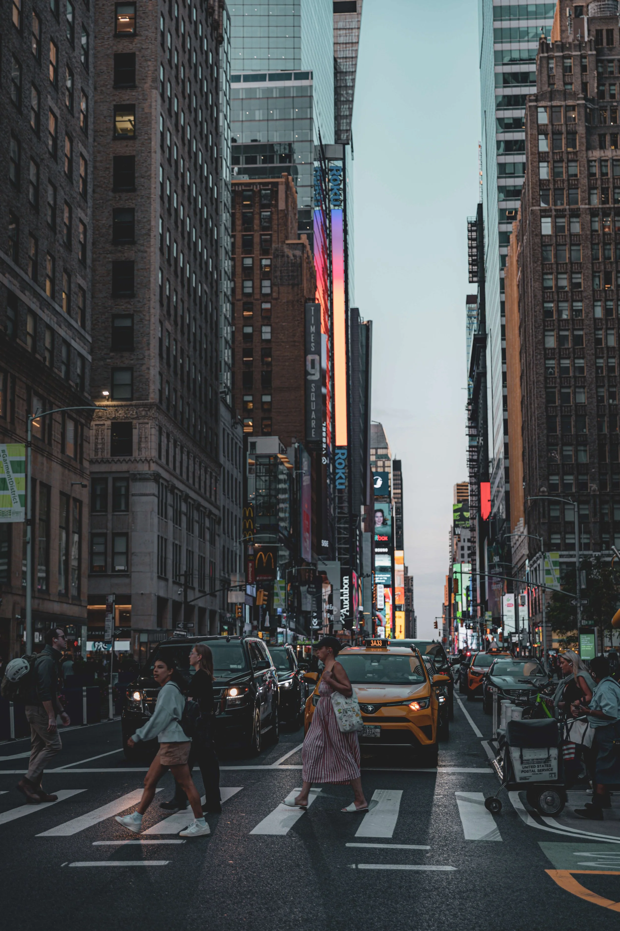 A busy city street in New York City with pedestrians crossing at a crosswalk, yellow taxis, and tall buildings with digital billboards and signs during dusk.