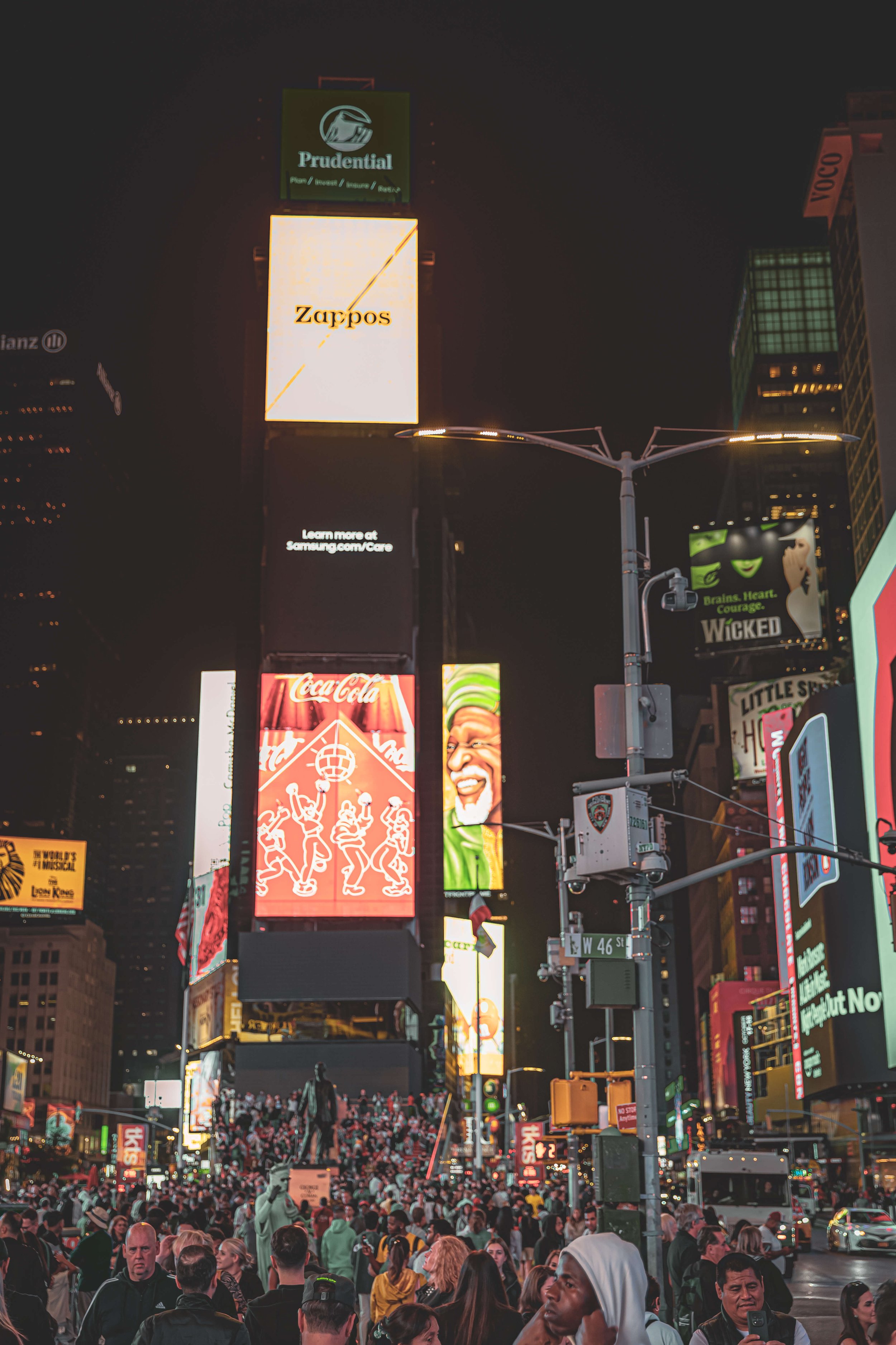 Nighttime scene in Times Square, New York City, with large digital billboards, a crowd of people, and illuminated advertisements.