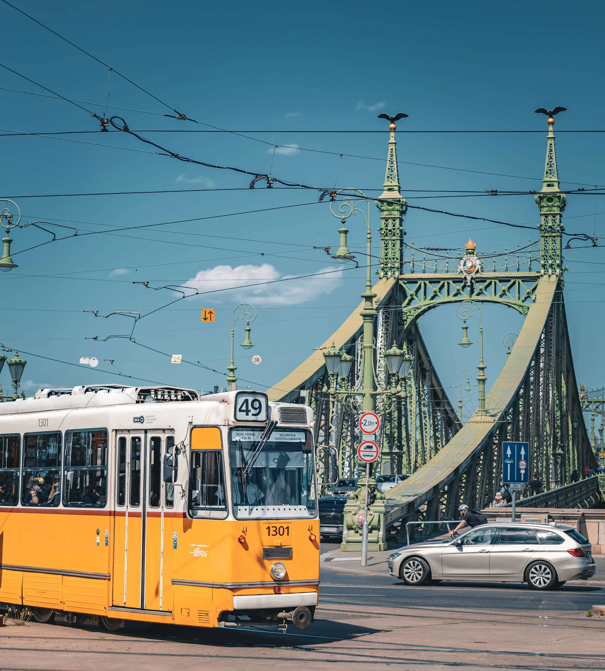 A yellow tram on the street with a city bridge in the background and overhead power lines crossing the sky, with a few cars and a scooter nearby.