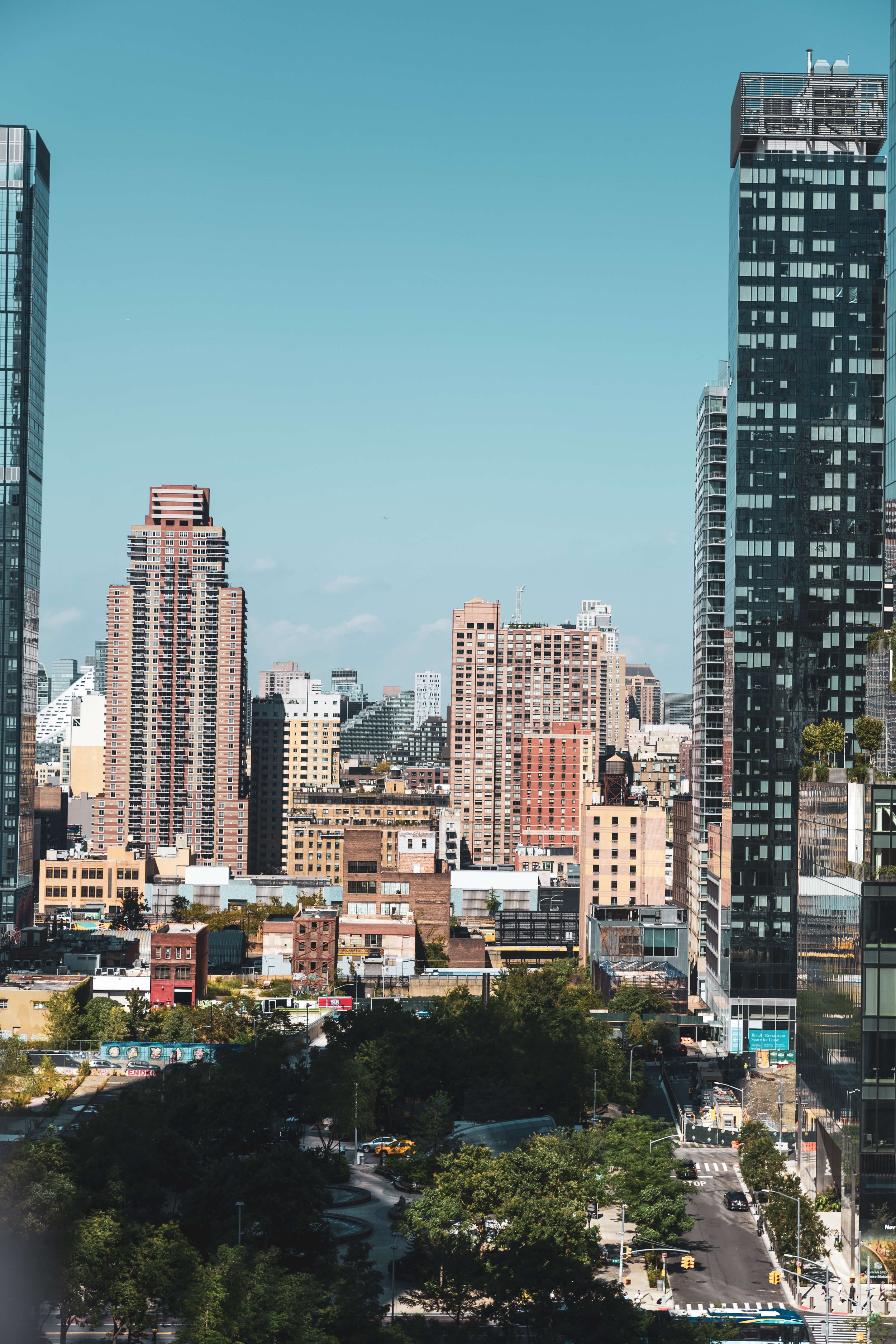City skyline with high-rise buildings under a clear blue sky, featuring both modern and older architecture, along with a street with cars and green trees in the foreground.