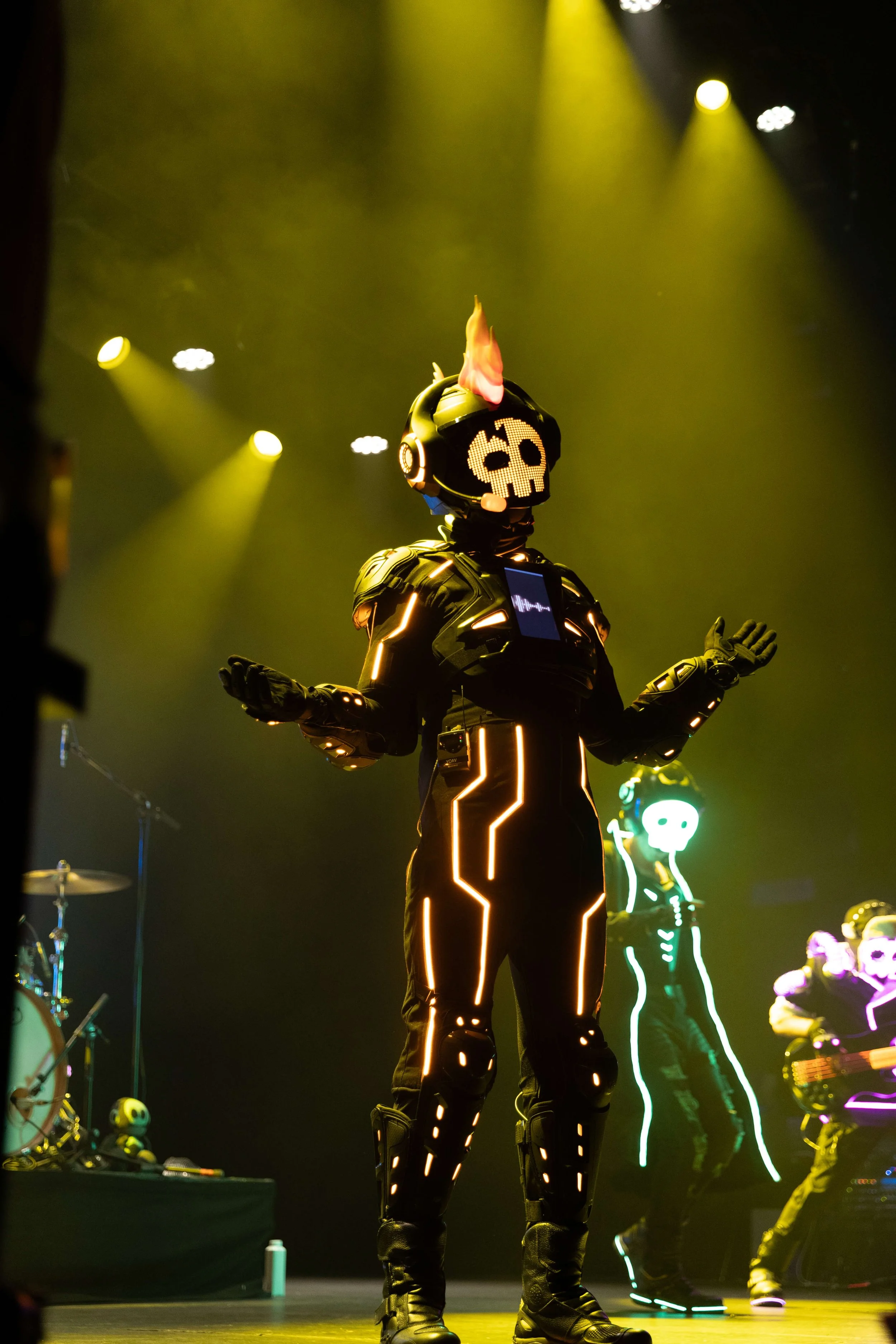A performer in a black costume with glowing neon lines on the suit, wearing a helmet with a skull LED face, a flaming mohawk, and headphones, stands with arms outstretched on stage with yellow and green lighting, surrounded by musicians with neon-lit