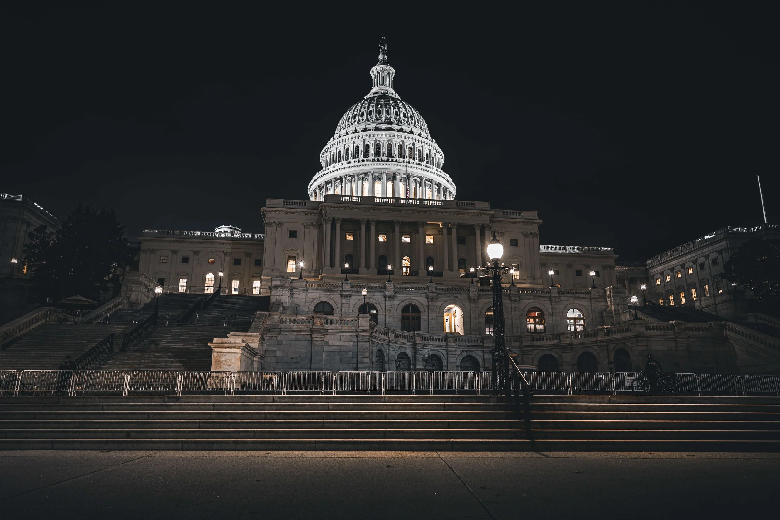 Nighttime view of the United States Capitol building with lit windows and illuminated dome, front steps, street lamps, and a fence in the foreground.