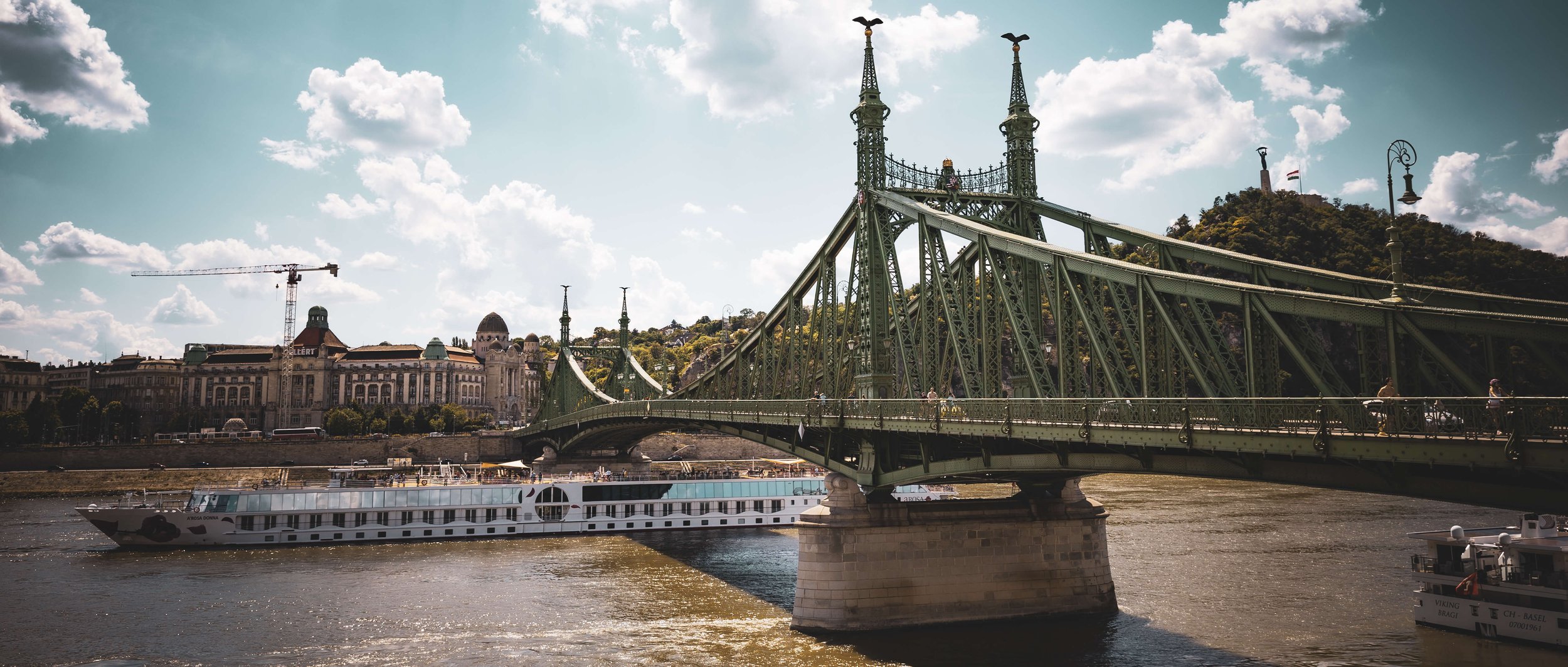 View of a green bridge crossing a river, with buildings and a hill in the background, and a boat sailing on the river.