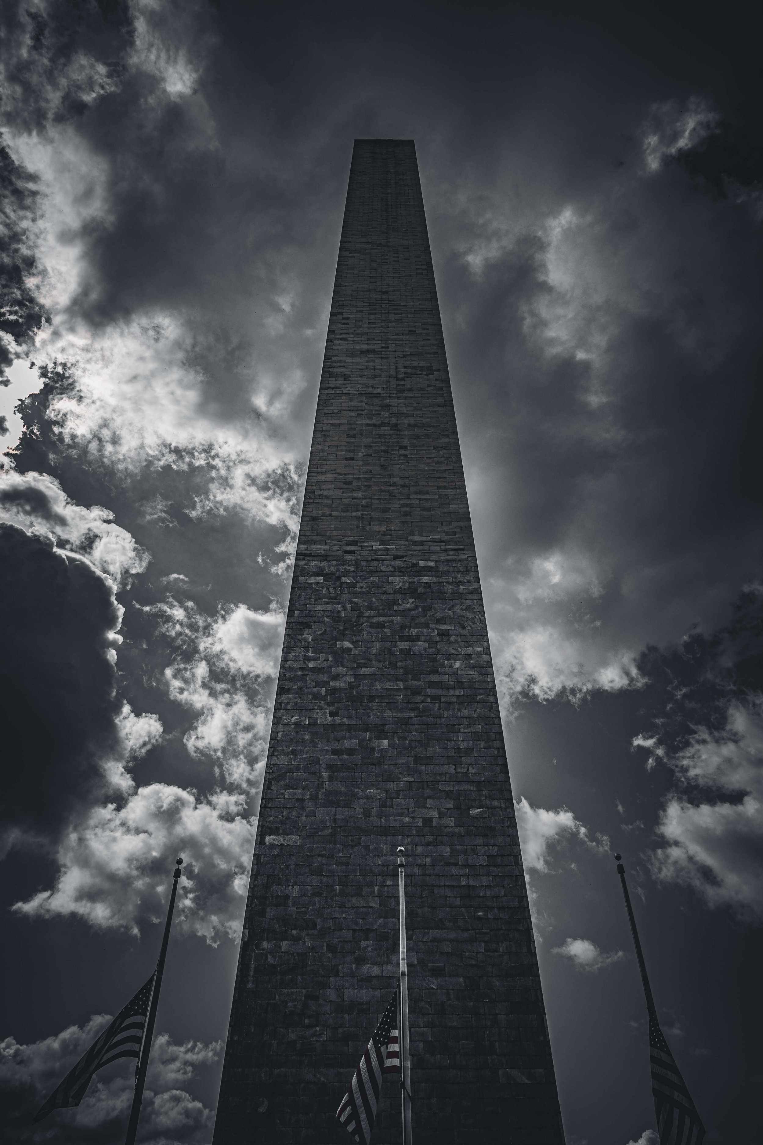 A tall stone obelisk monument against a cloudy sky, with four American flags at its base.