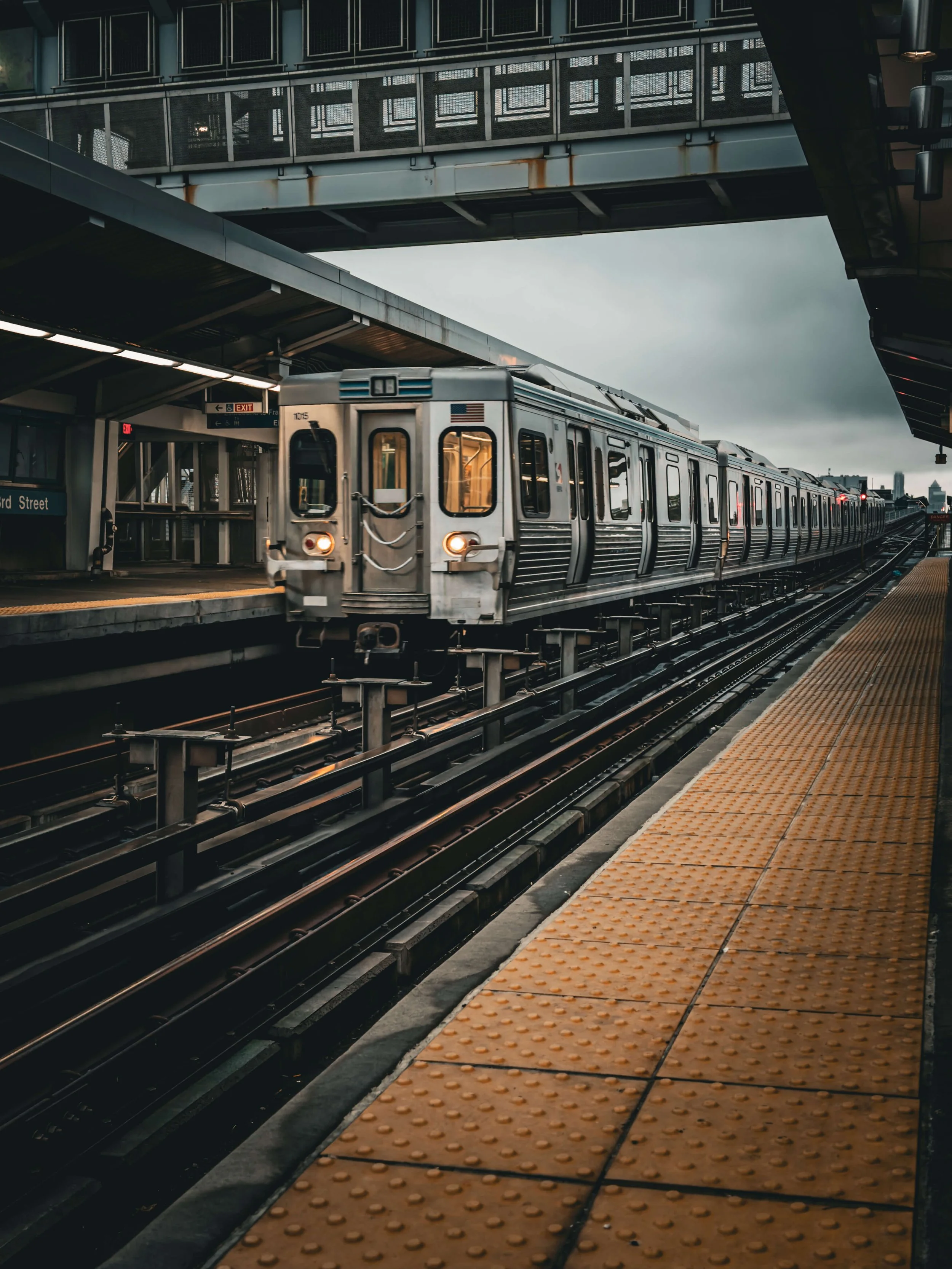 A city subway train arriving at an outdoor station on a cloudy day, with overhead walkways and platform safety markings.