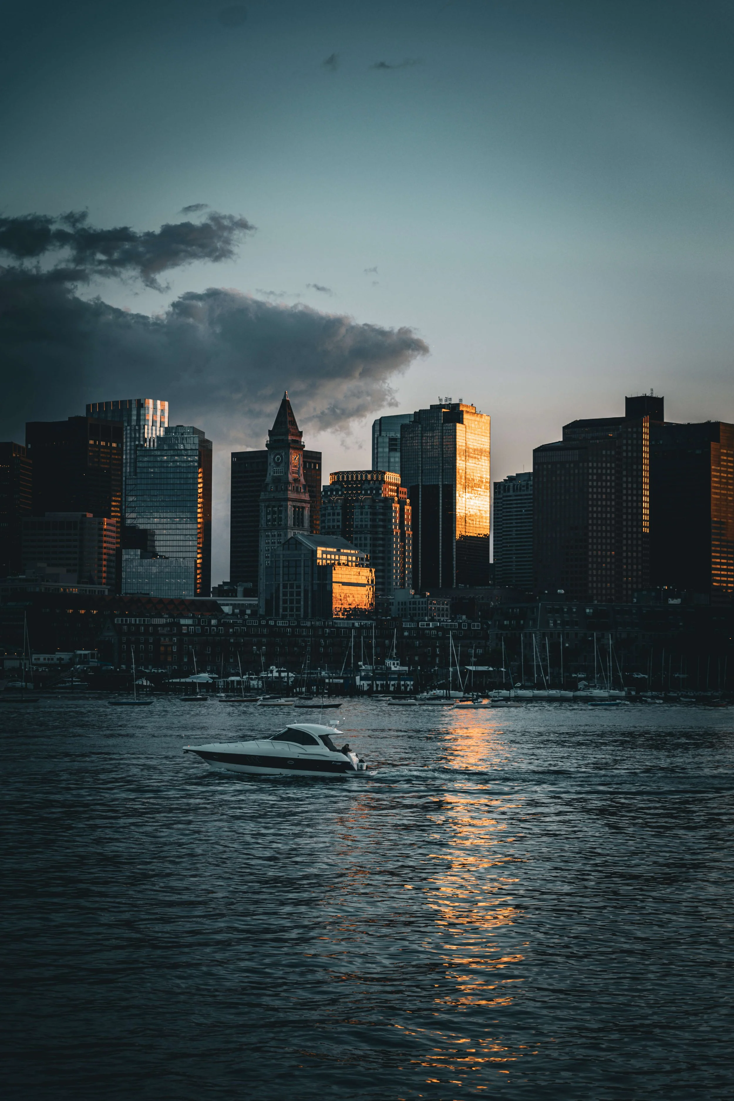 A city skyline with tall buildings reflected in the water, a boat in the foreground, and a cloudy sky at dusk.