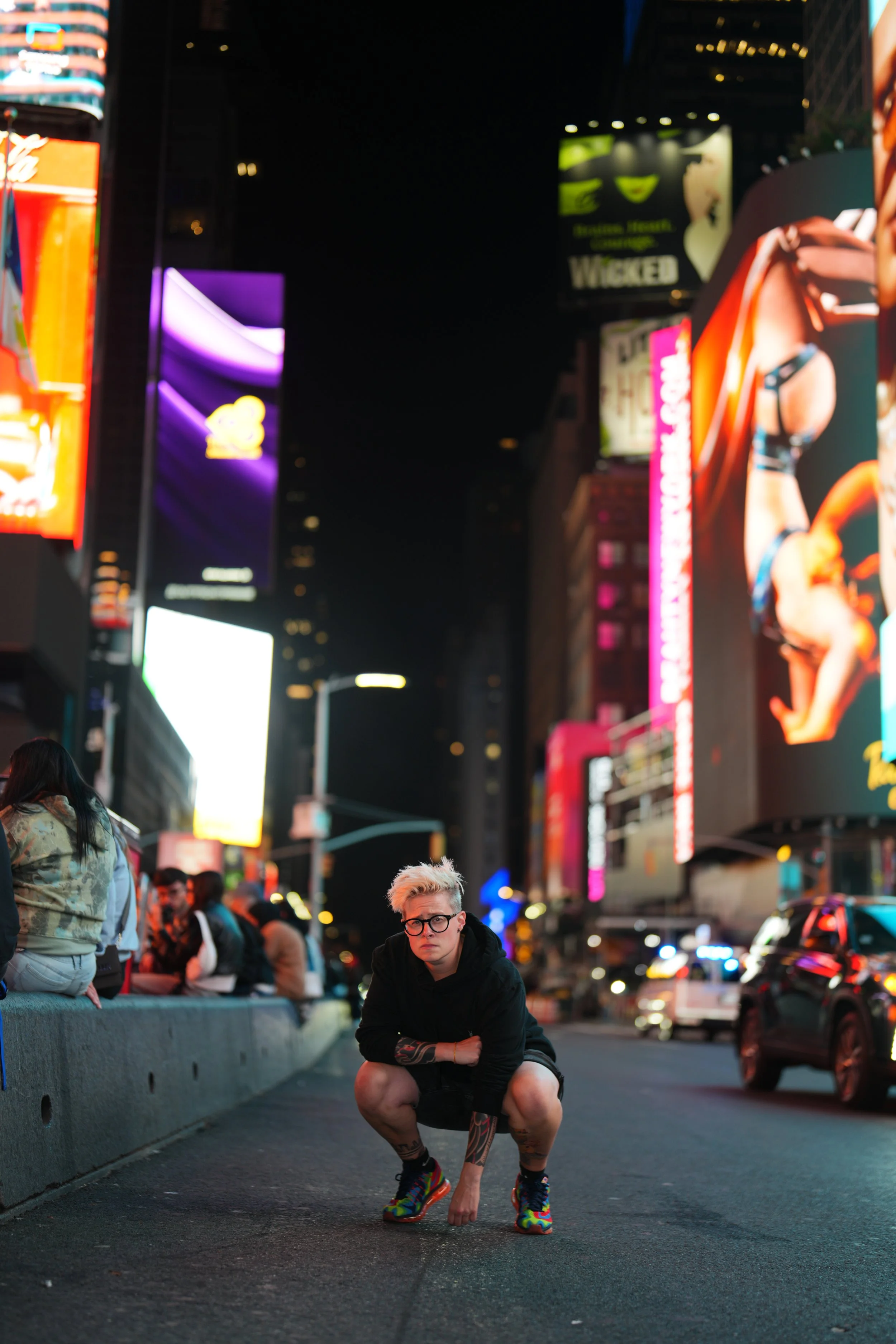 A young woman with tattoos, glasses, white spiked hair and colorful shoes crouches on a city street at night, surrounded by neon billboards and other pedestrians.