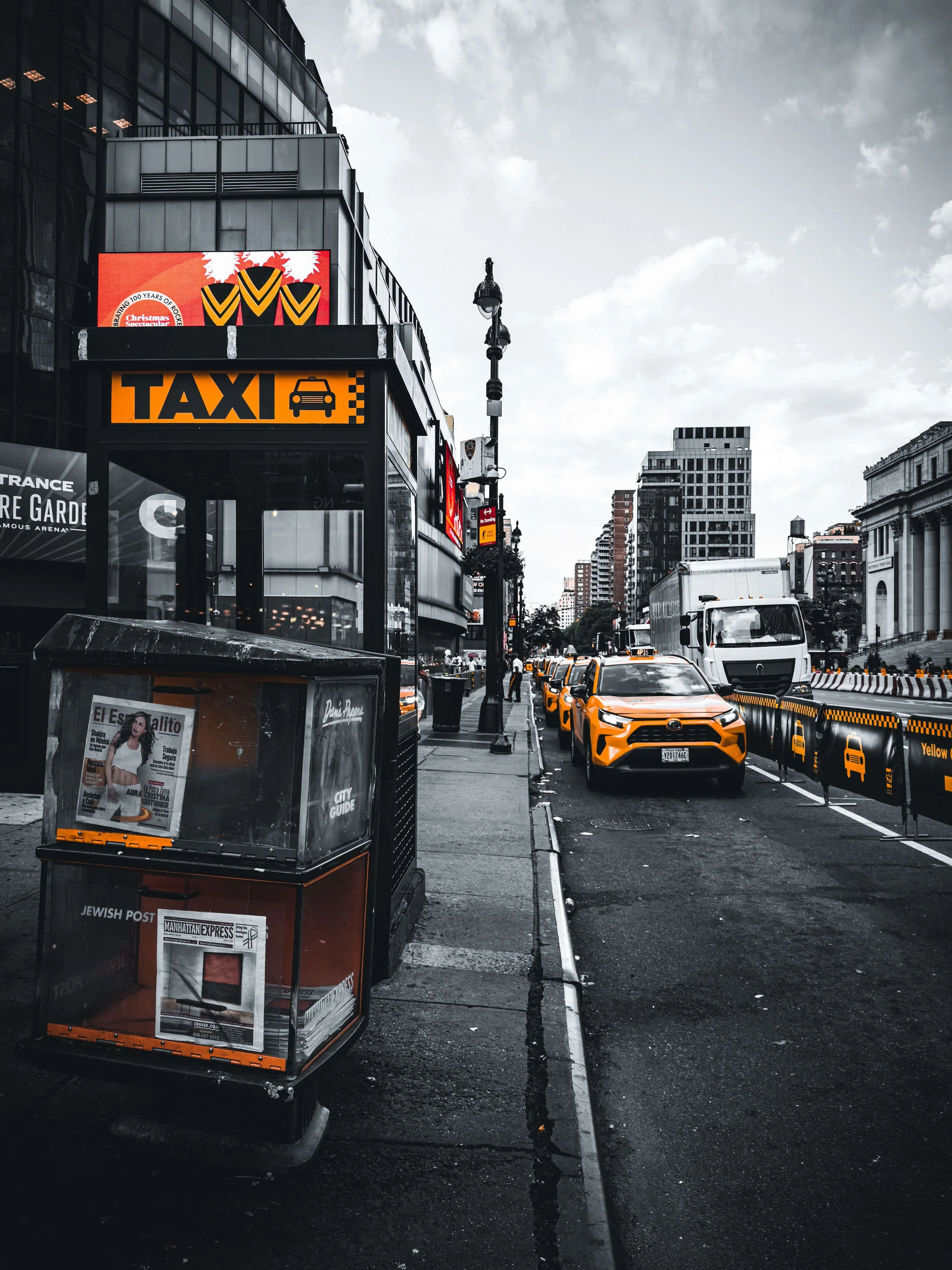 City street scene with taxi stand, lined with yellow taxis and other vehicles, buildings, and a newspaper box in the foreground.