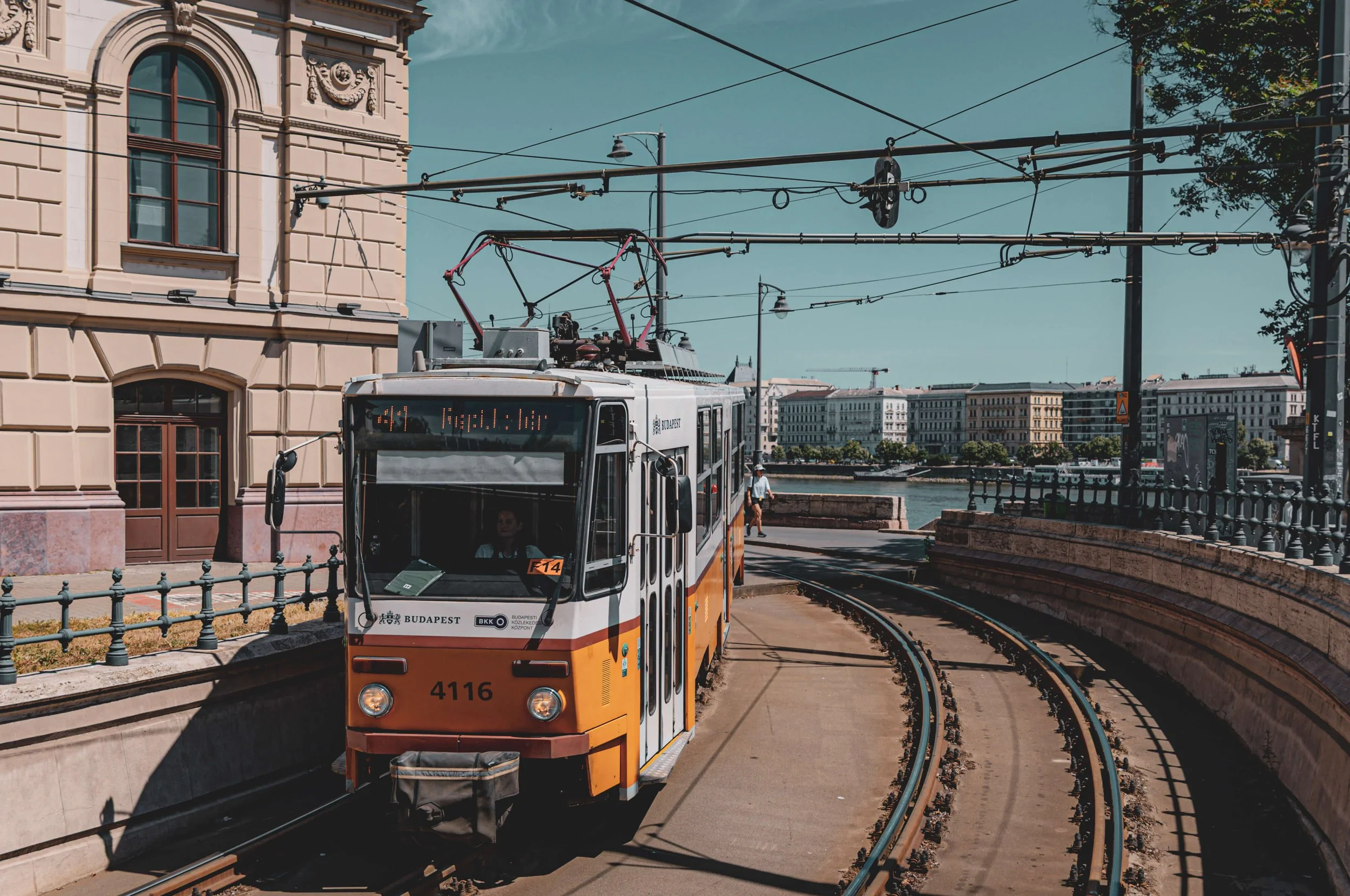 A tram with orange, white, and black colors traveling on curved tracks along a city street in Budapest, Hungary. The tram displays a route sign, and there are historic buildings and infrastructure with overhead wires for the tram.
