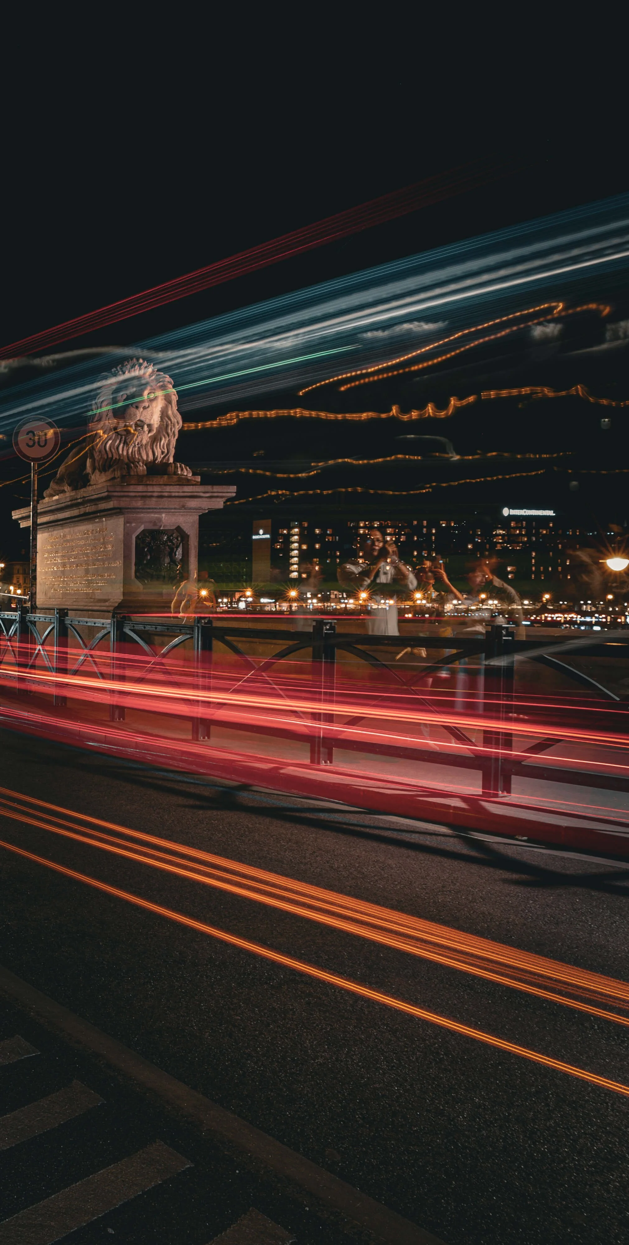 Night scene of a historic stone lion statue on a city bridge with colorful light streaks from moving cars and city lights in the background.