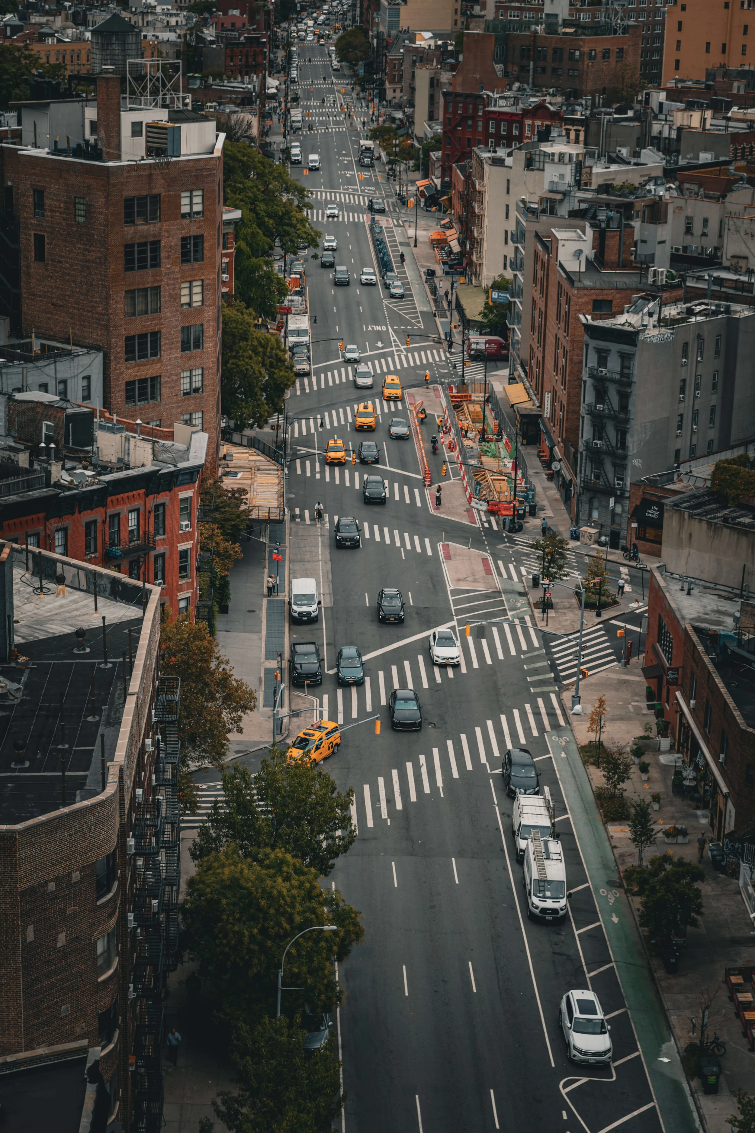 Aerial view of a busy city street with cars, taxis, and pedestrians, lined with brick and concrete buildings and trees.