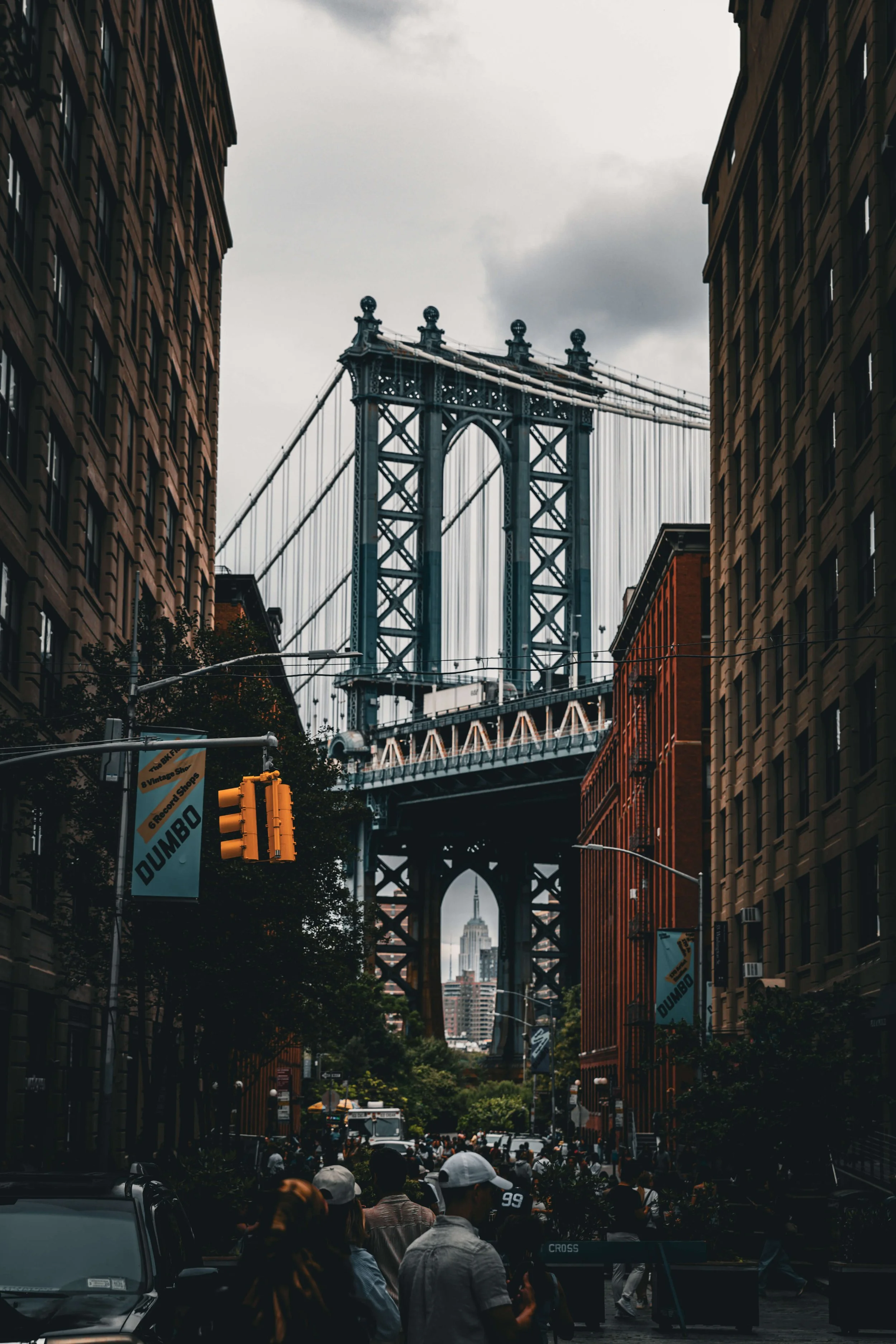 People walking in a busy city street with the Manhattan Bridge in the background, and the Empire State Building visible through the bridge's arch.