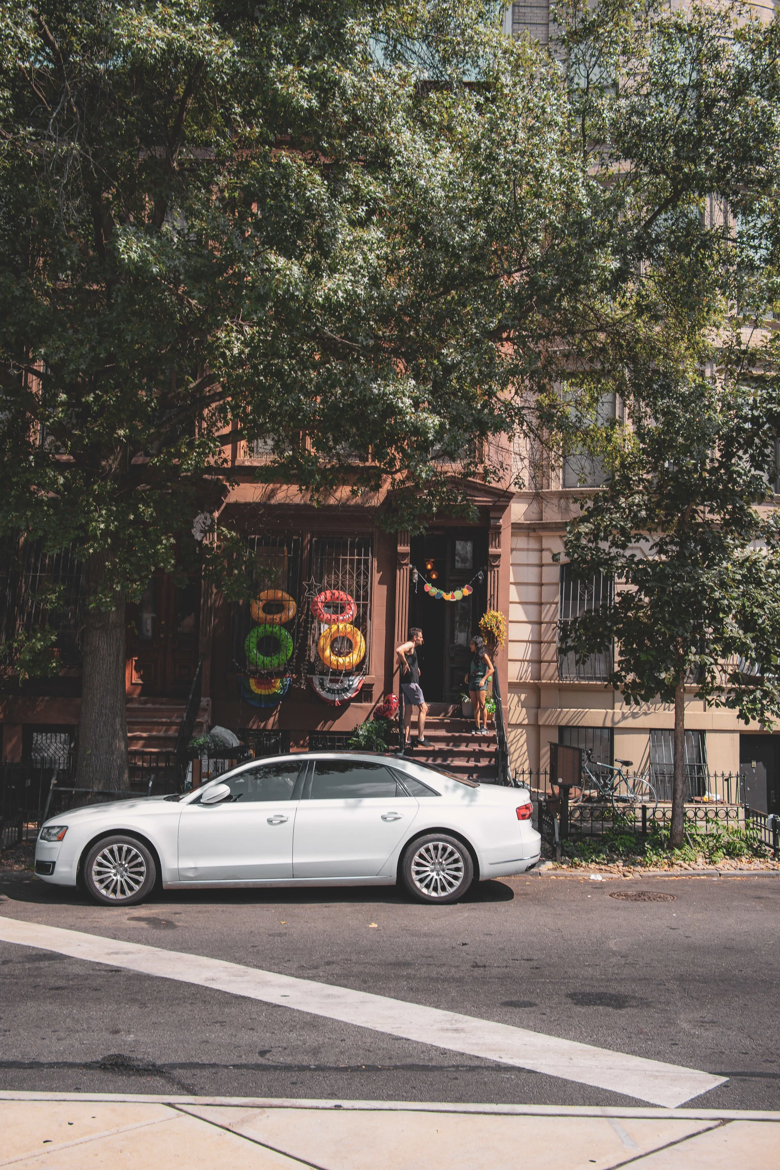 People are standing on stairs decorated with colorful float rings and garlands outside a brownstone building, with a white car parked on the street in front.