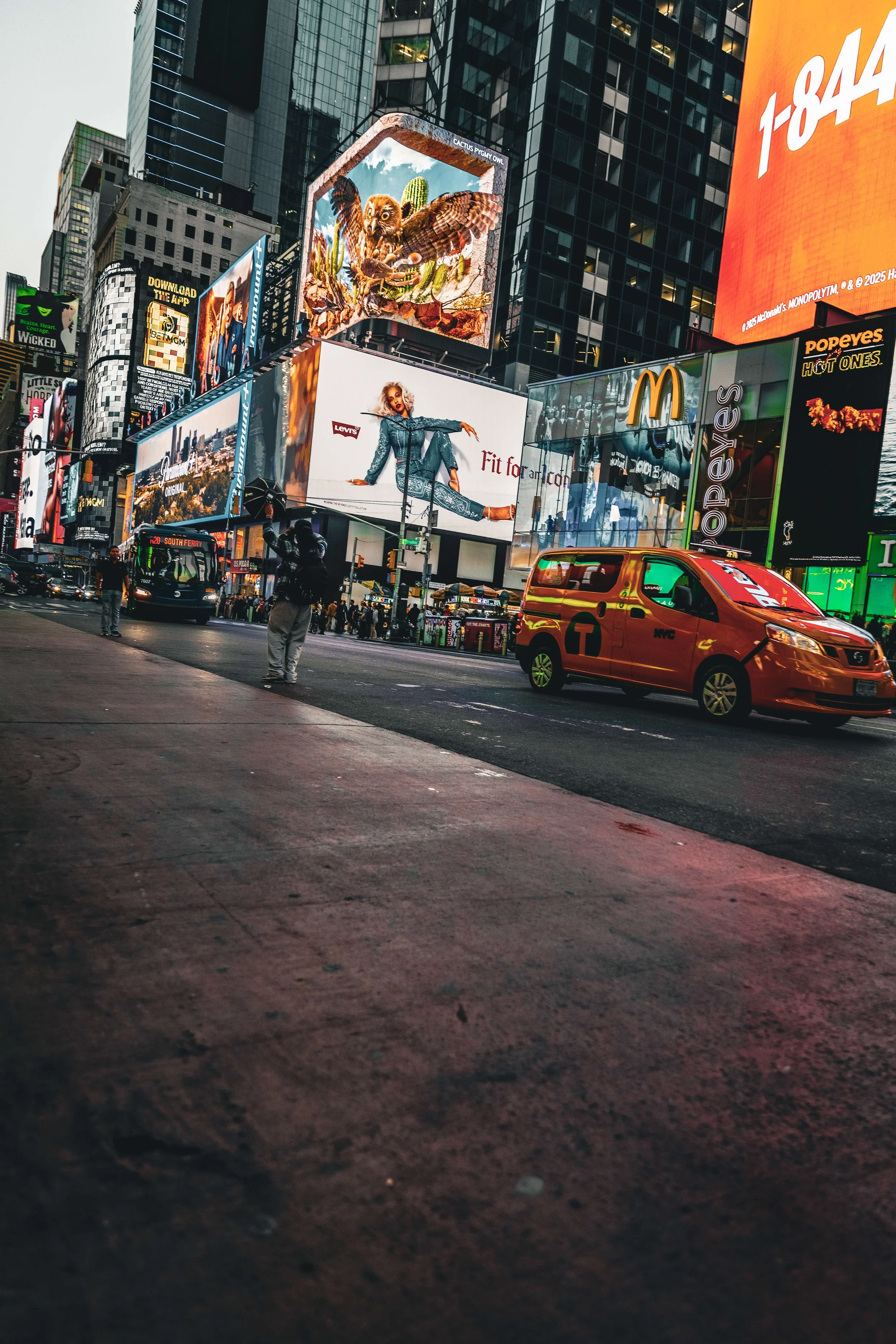 City street scene in Times Square, New York City, featuring large digital billboards advertising fashion, entertainment, and food. There is a taxi and a bus on the street, with a few pedestrians on the sidewalk.