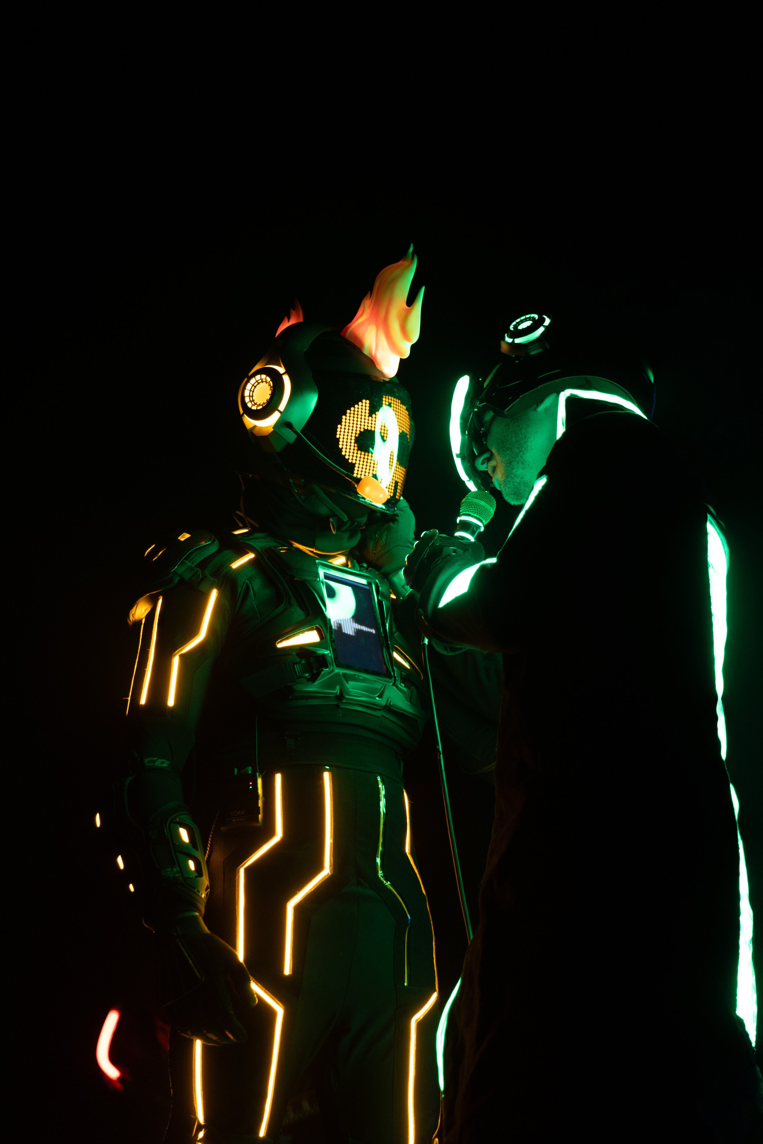 Two people dressed in futuristic costumes with glowing LED lights and masks, standing in darkness. One person has a helmet with a digital display, and the other wears a mask with flames and a digital dollar sign.