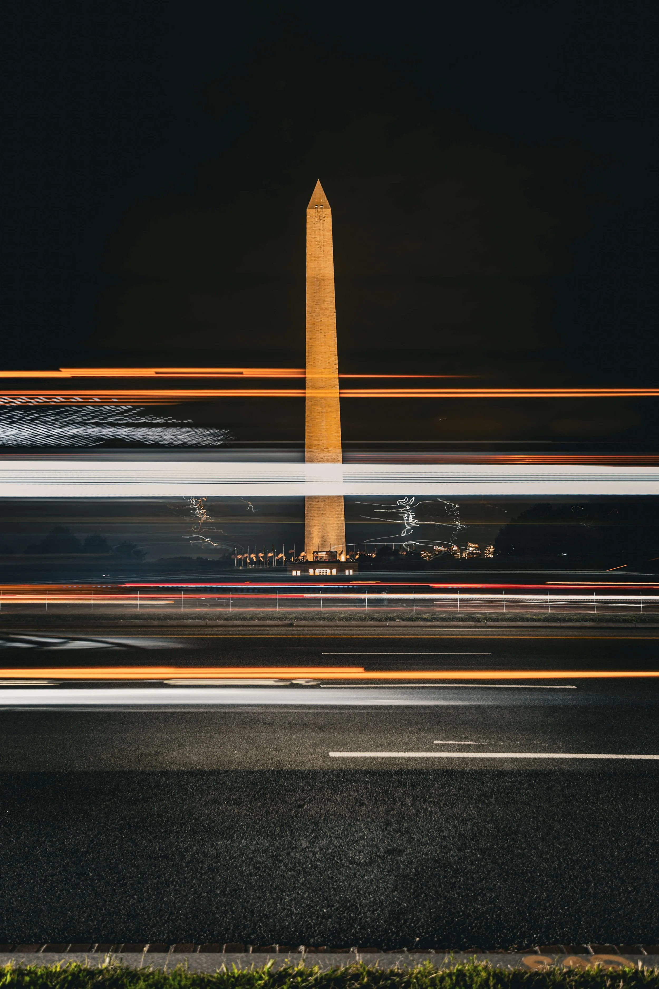 Long exposure nighttime photo of the Washington Monument with light trails from passing vehicles in the foreground.