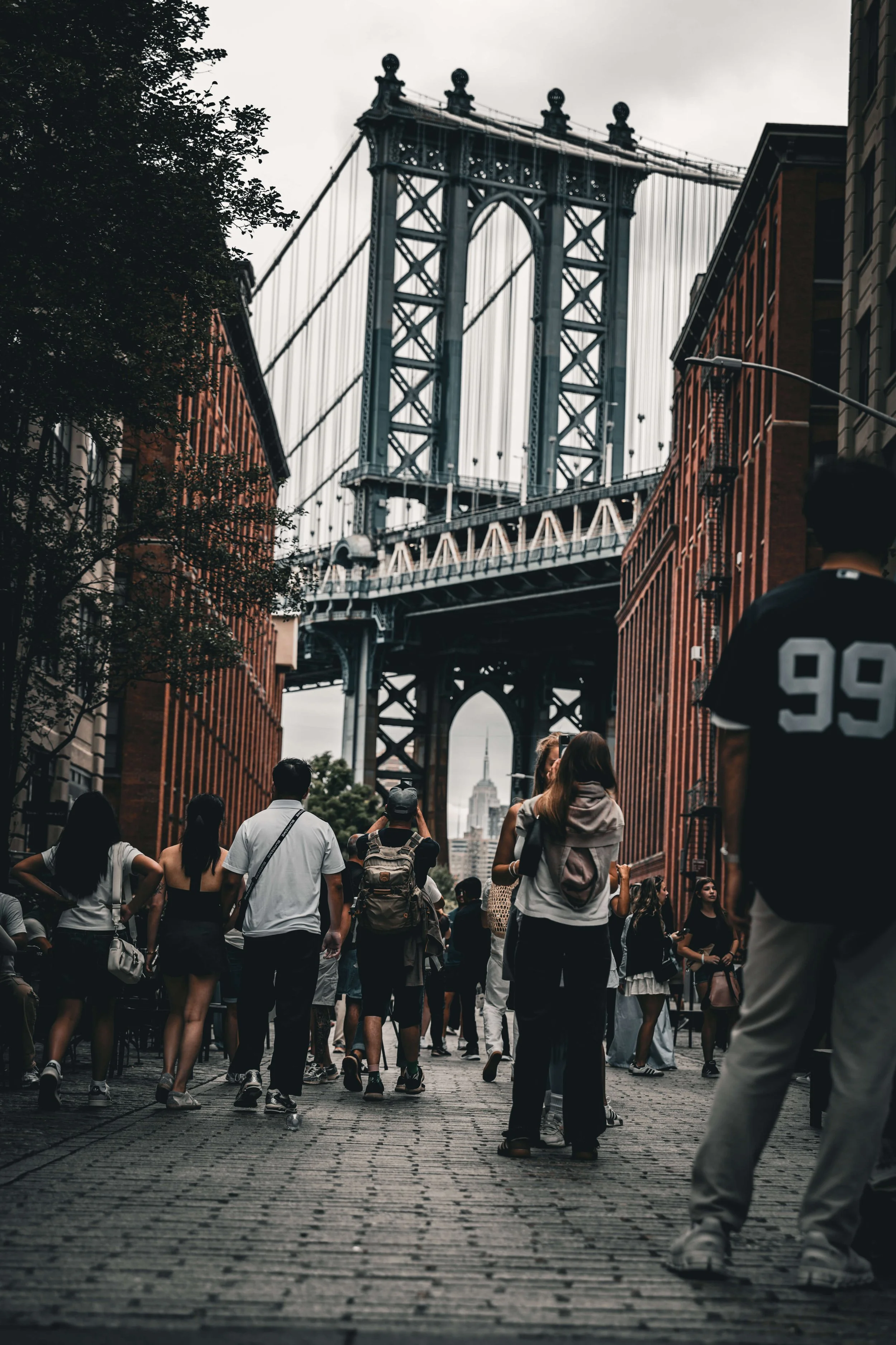 Crowd of people walking on cobblestone street with Brooklyn Bridge and Empire State Building in background, overcast sky.