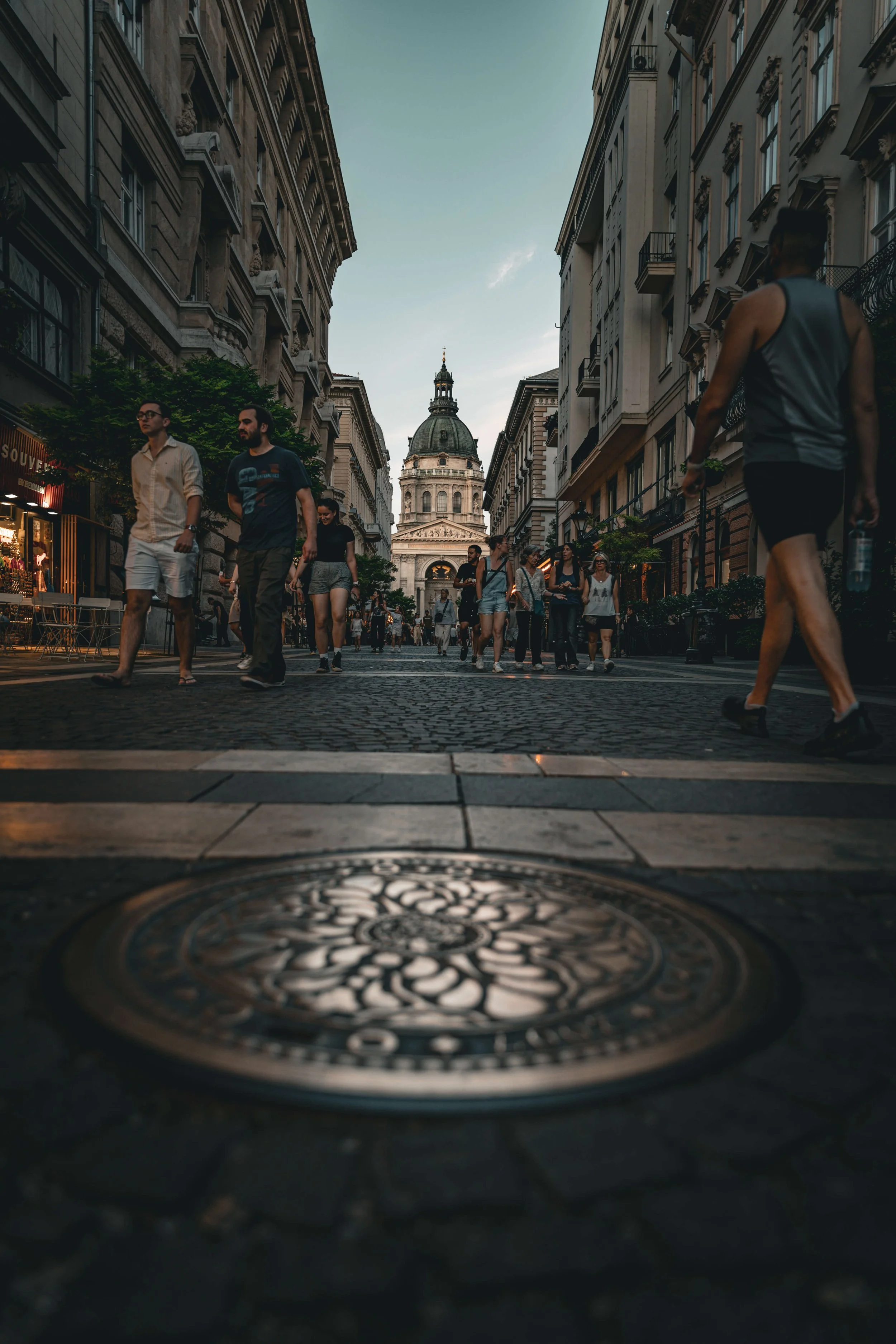 People walking on a city street with historic buildings and a domed church in the background, viewed from low angle.