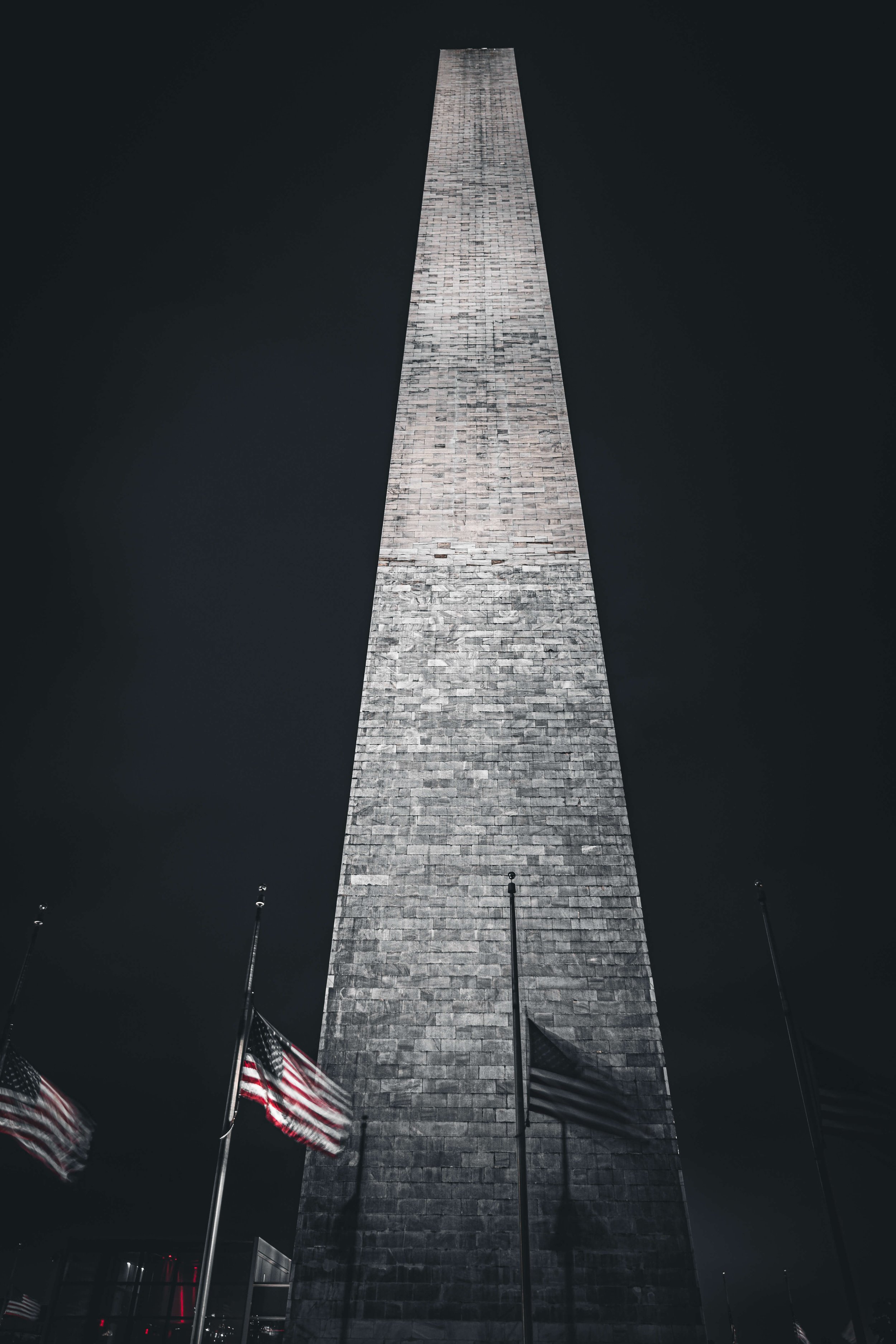 A tall, illuminated stone obelisk monument at night with American flags in front of it against a dark sky.