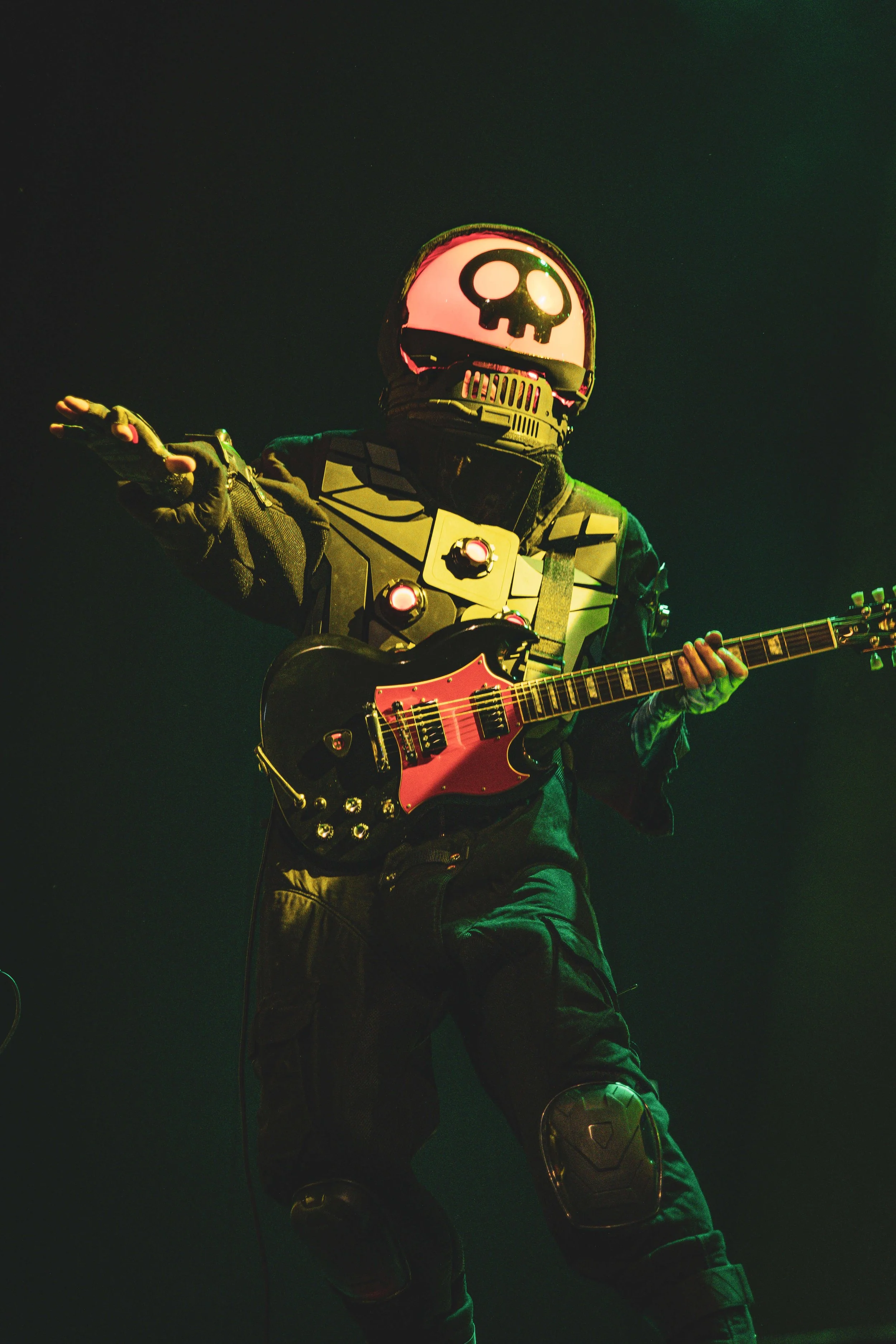 A person dressed as a superhero with a skull logo on their helmet, wearing a futuristic suit with armor plates, playing an electric guitar on stage with dark lighting and greenish hues.